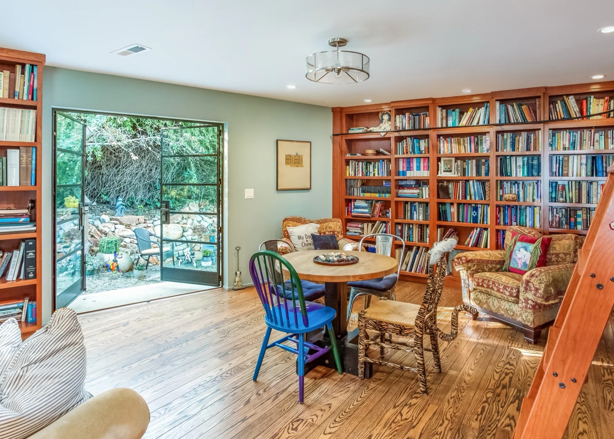 Living room with wooden floors, large bookshelf filled with books, a round wooden table with colorful chairs, a floral armchair, and an open glass door leading to a garden with rocks and trees.