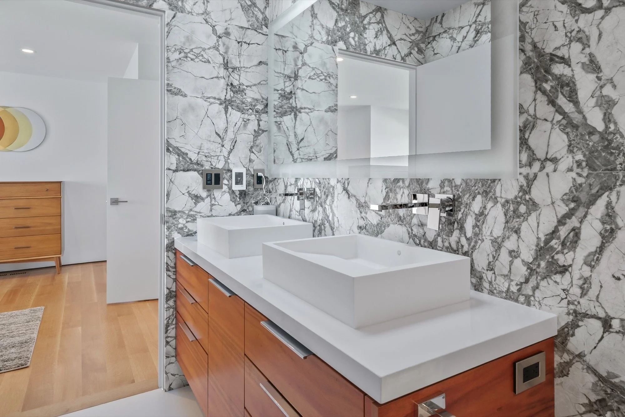Modern bathroom with marble walls and two rectangular vessel sinks on a white countertop, wooden vanity, and large mirror.