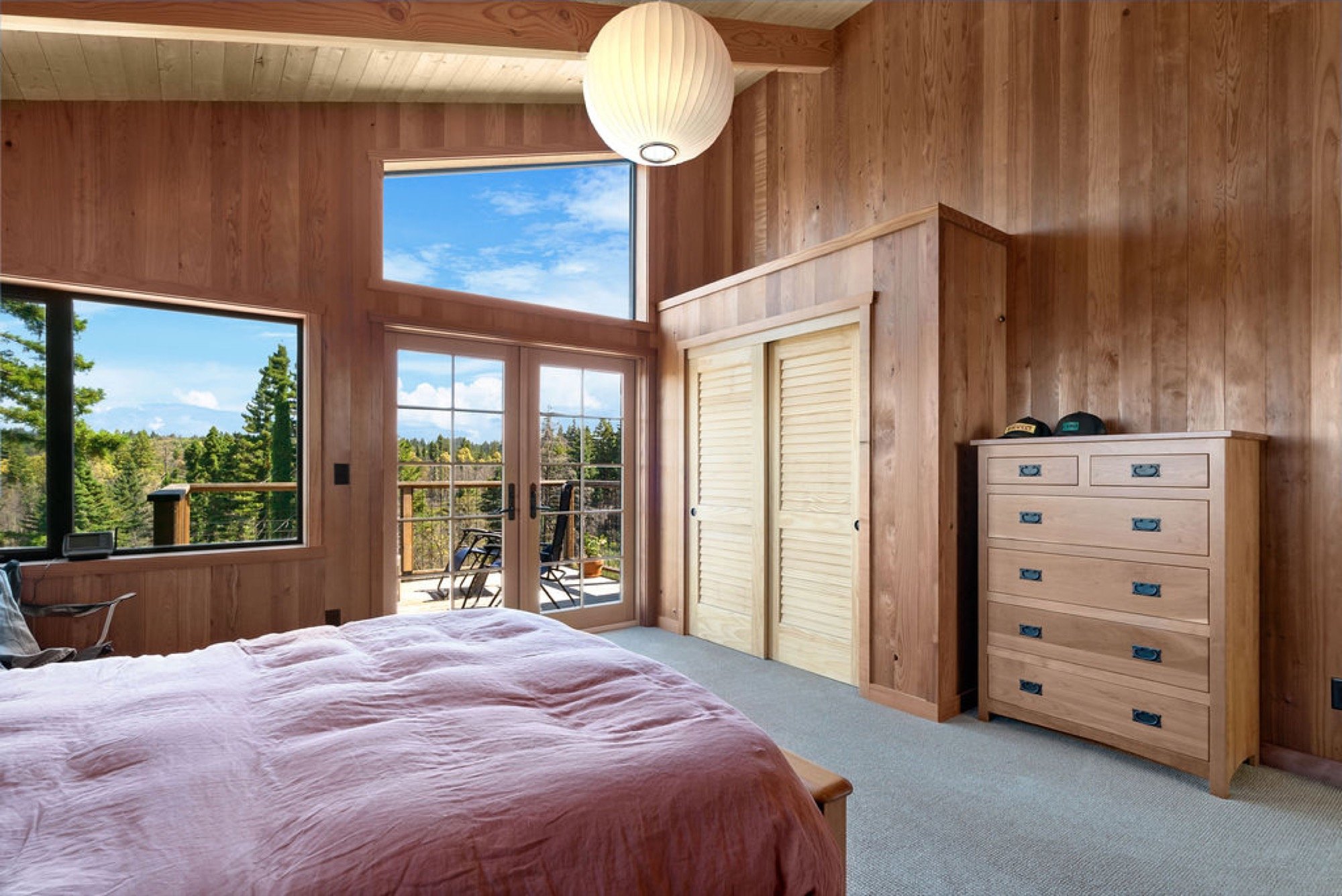 A bedroom with wooden walls, a bed with a pink blanket, a dresser with six drawers, and a sliding glass door leading to a balcony with outdoor chairs. There are large windows showing a view of trees and sky.