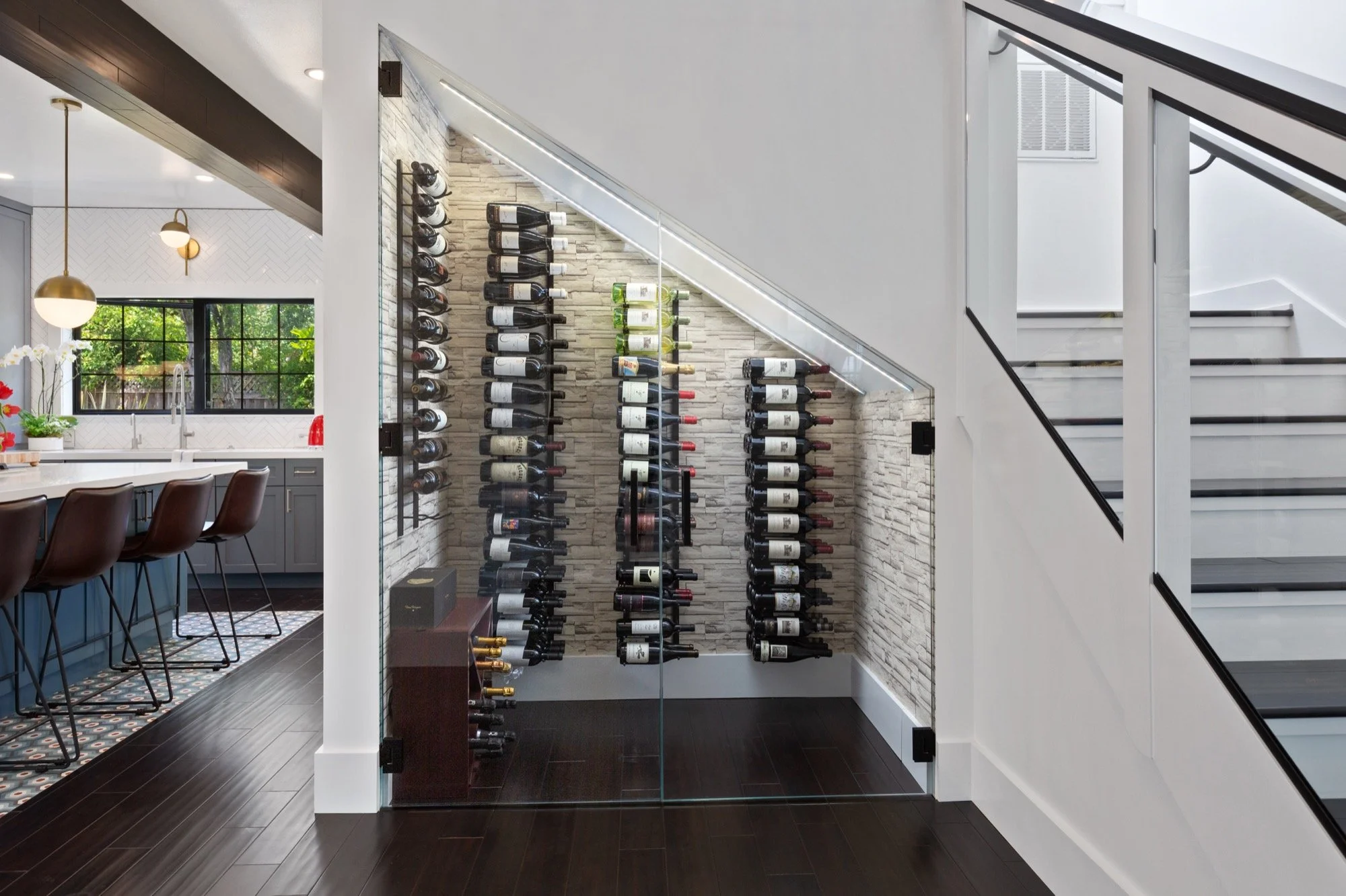 Modern interior of a home with a wine storage area under a staircase, featuring a glass wall and wine bottles organized on wall-mounted racks.