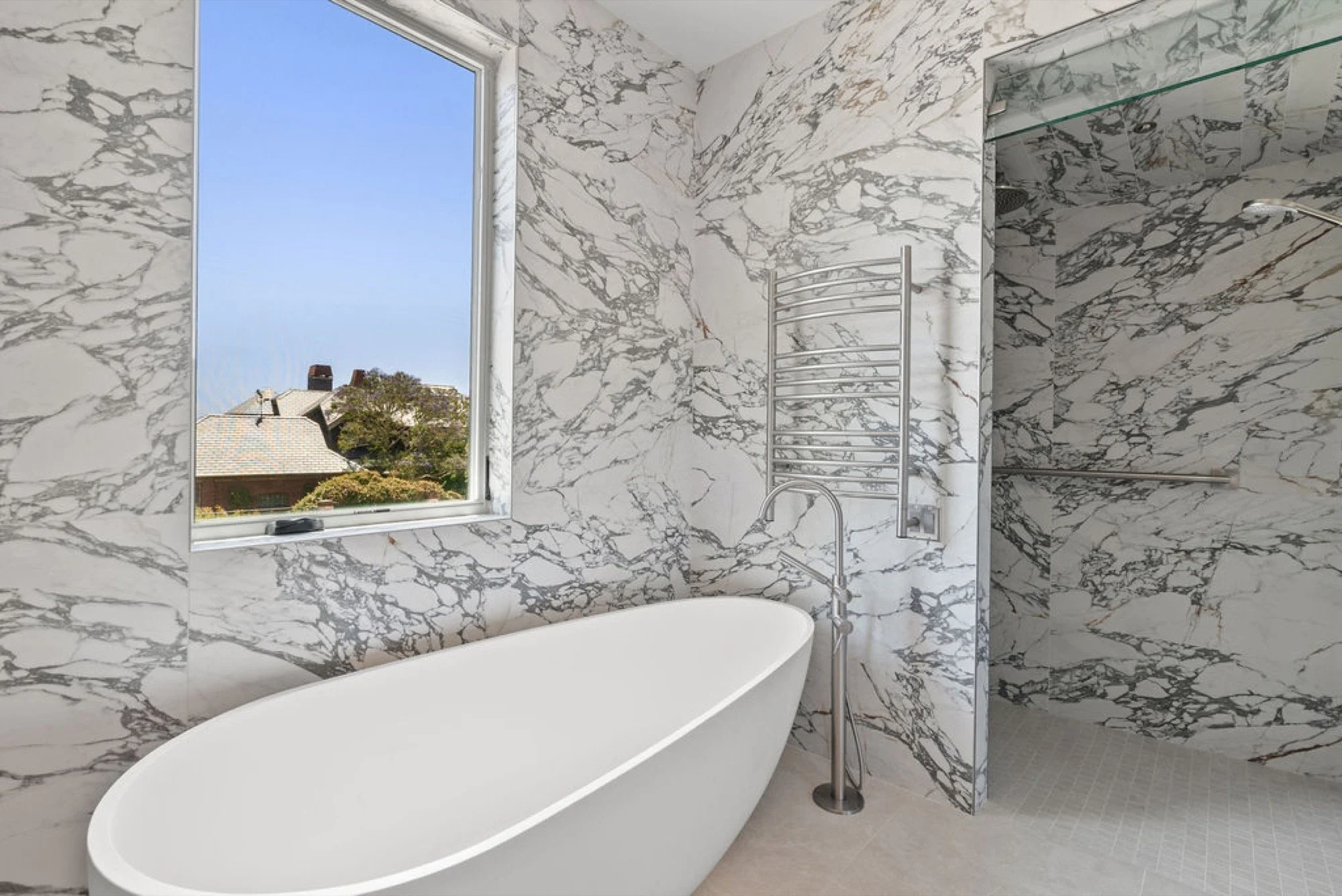 A modern bathroom featuring a white freestanding bathtub, marble walls with gray veining, a window showing a view of rooftops and trees, and a chrome towel warmer.
