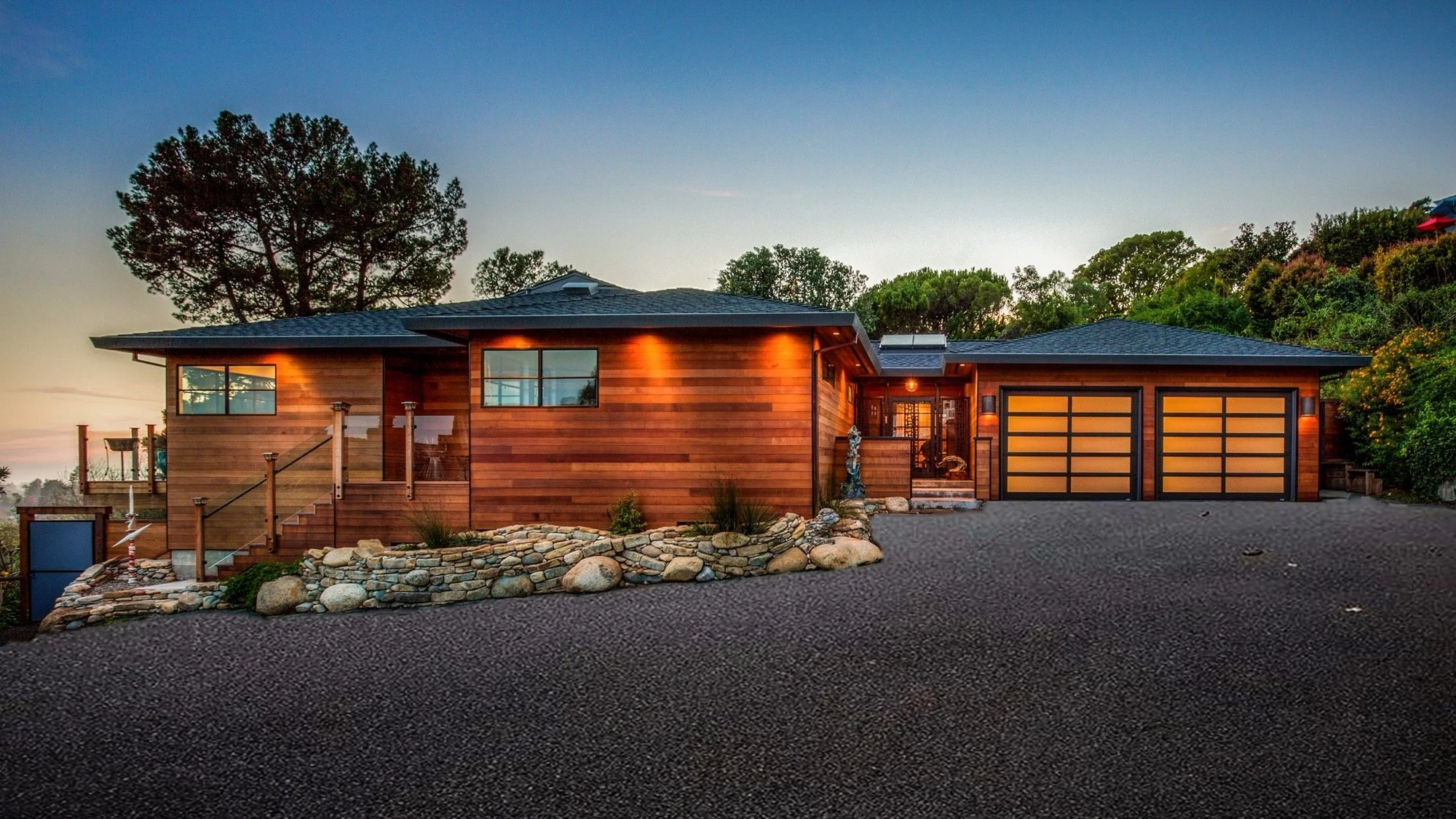 Modern house with wooden siding, stone landscaping, and a double garage, set against a backdrop of trees during sunset.