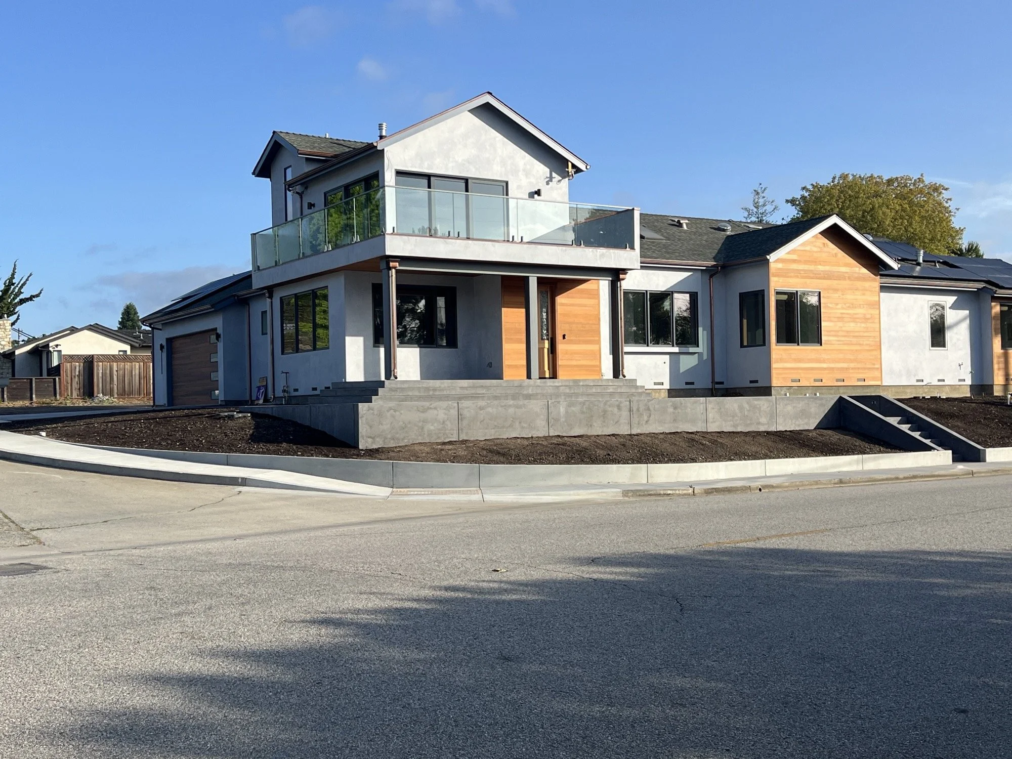 A modern two-story house with white walls, wooden accents, large windows, a balcony, and a garage on a corner lot in a suburban neighborhood.