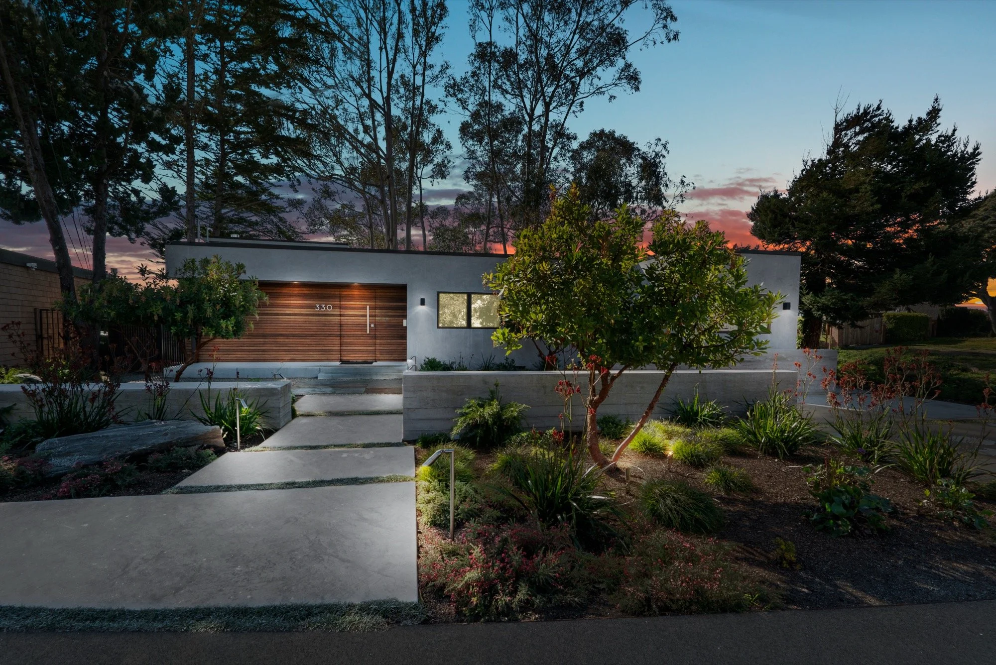 Modern single-story house with a wooden front door, large window, and concrete pathway leading to the entrance, surrounded by landscaped garden with trees and plants at sunset.