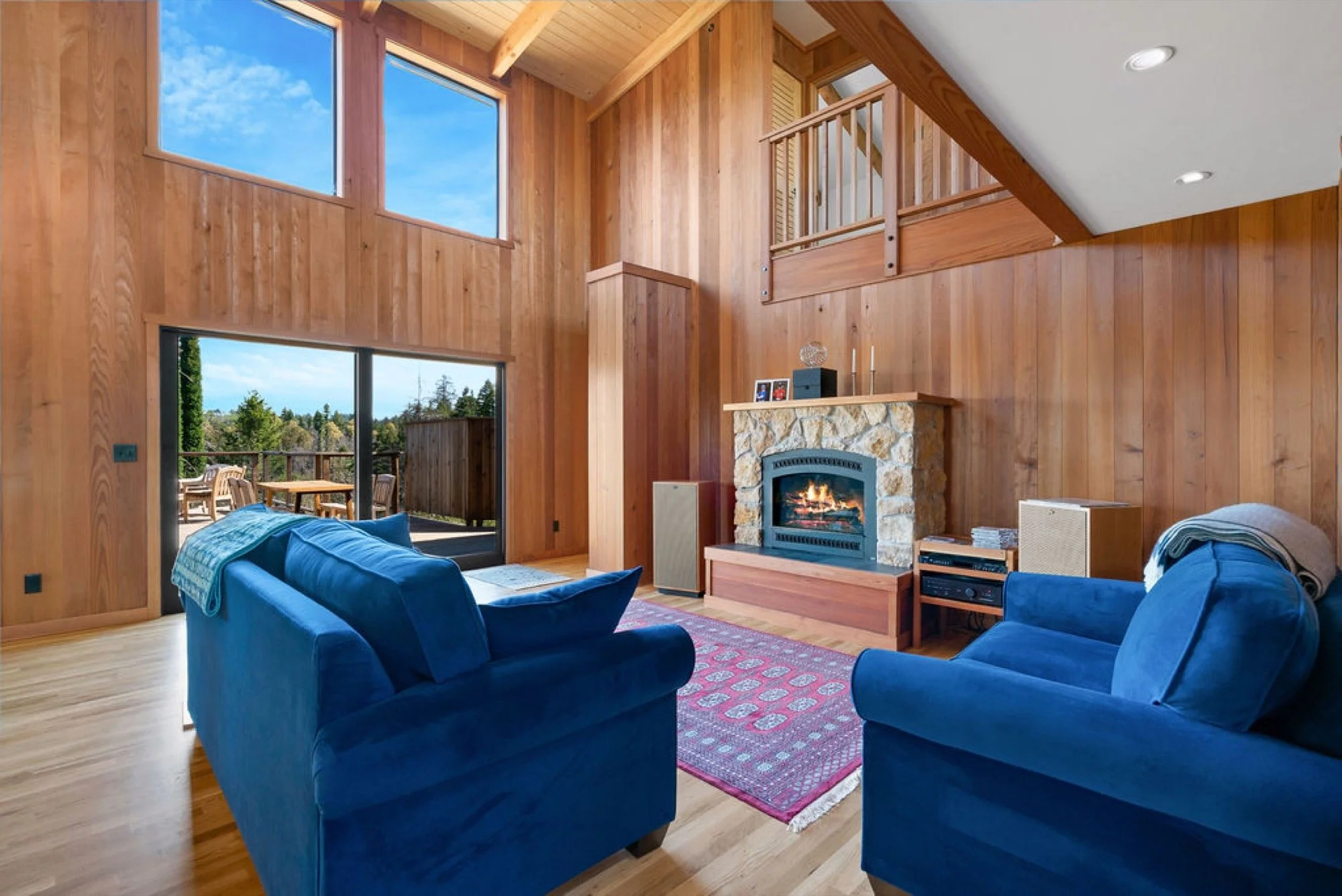 Living room with wooden walls and ceiling, two blue sofas, a stone fireplace with fire, a colorful area rug, large windows and sliding glass door leading to a balcony with outdoor furniture, and a staircase overhead.