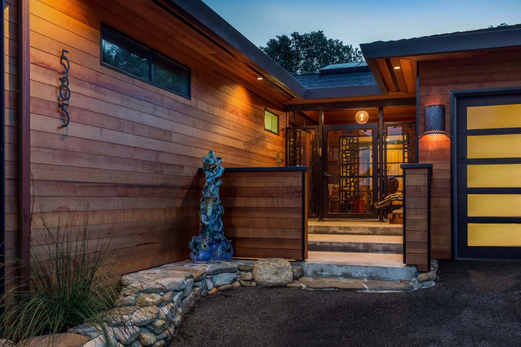 Front view of a modern house with wood exterior, steps leading to the entrance, decorative sculpture on the left, and warm exterior lighting at dusk