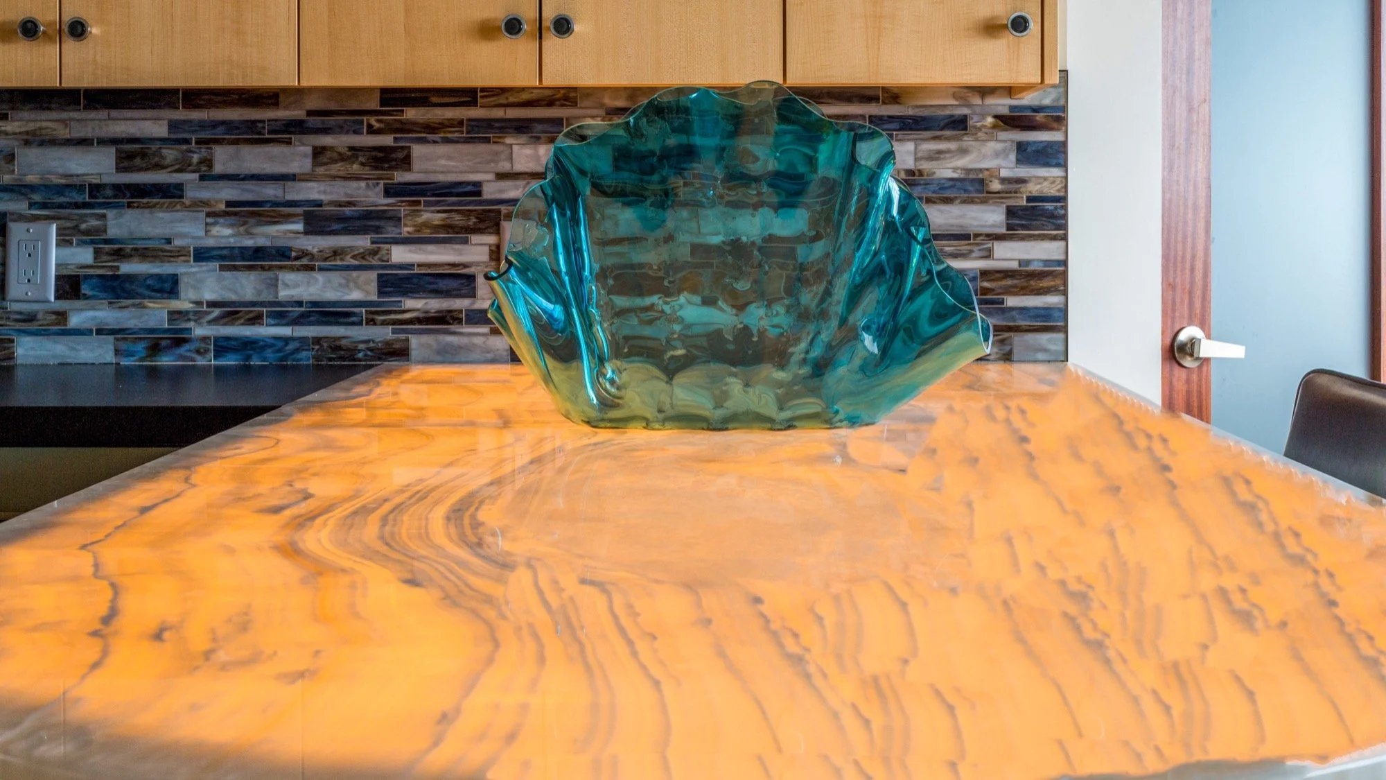 A glass art piece resembling a large, irregularly shaped shell or flower with blue and yellow-green hues, placed on a polished orange wood table in a modern kitchen or dining area.