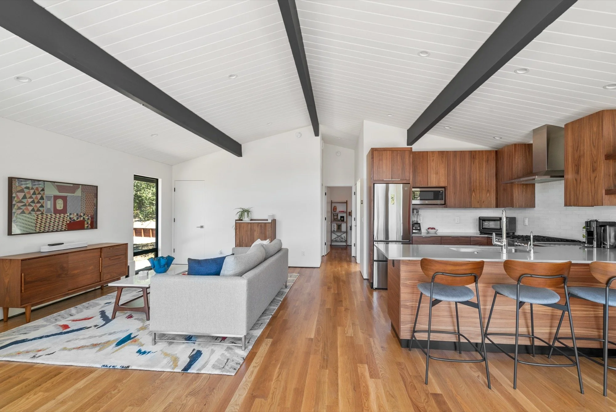 Open-concept living room and kitchen with white shiplap ceiling, wooden beams, hardwood flooring, gray sectional couch, colorful rug, wooden media console, modern kitchen with wooden cabinets, stainless steel appliances, and a breakfast bar with four