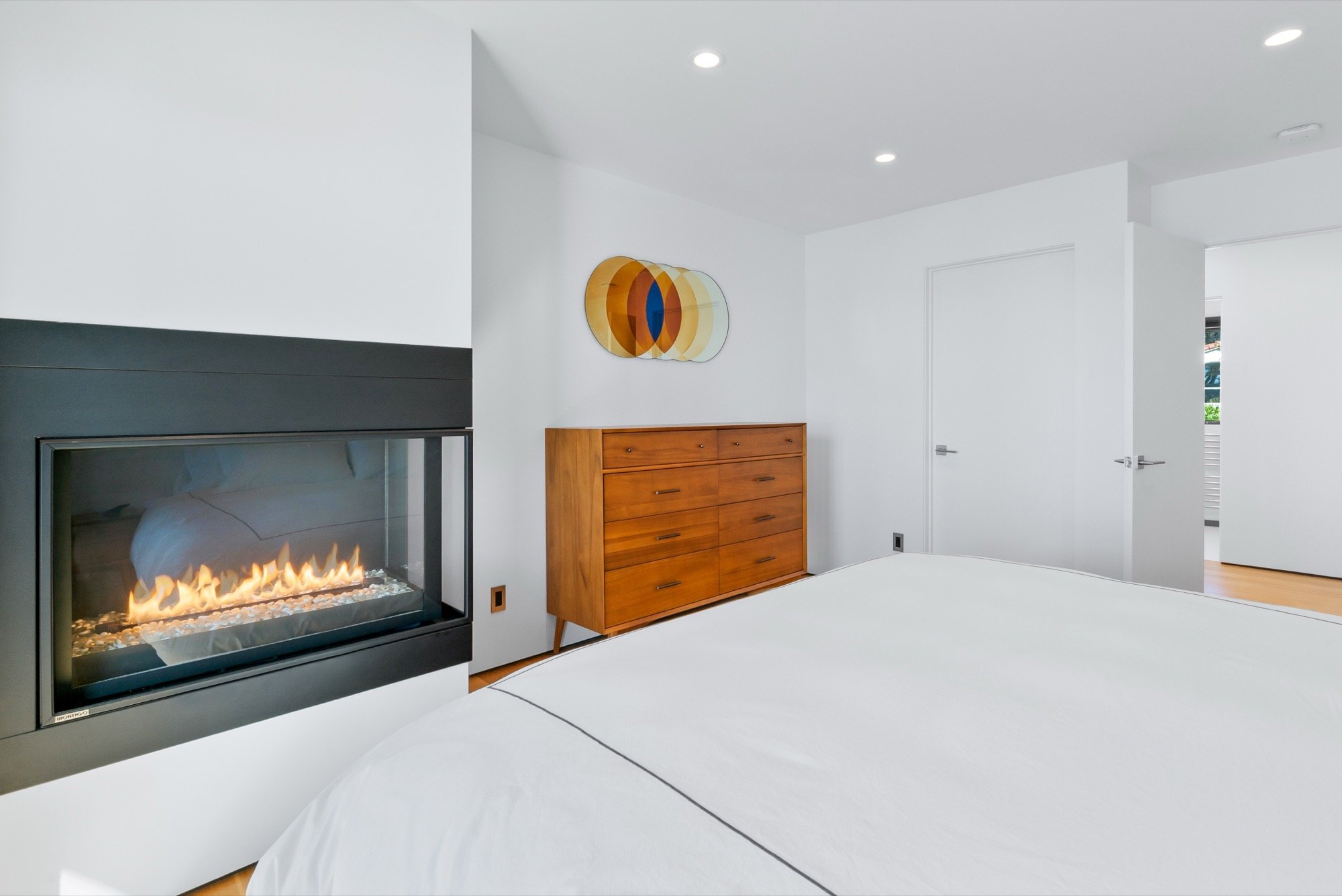 Modern bedroom with a white bed, a fireplace, a wooden dresser, and a wall art piece