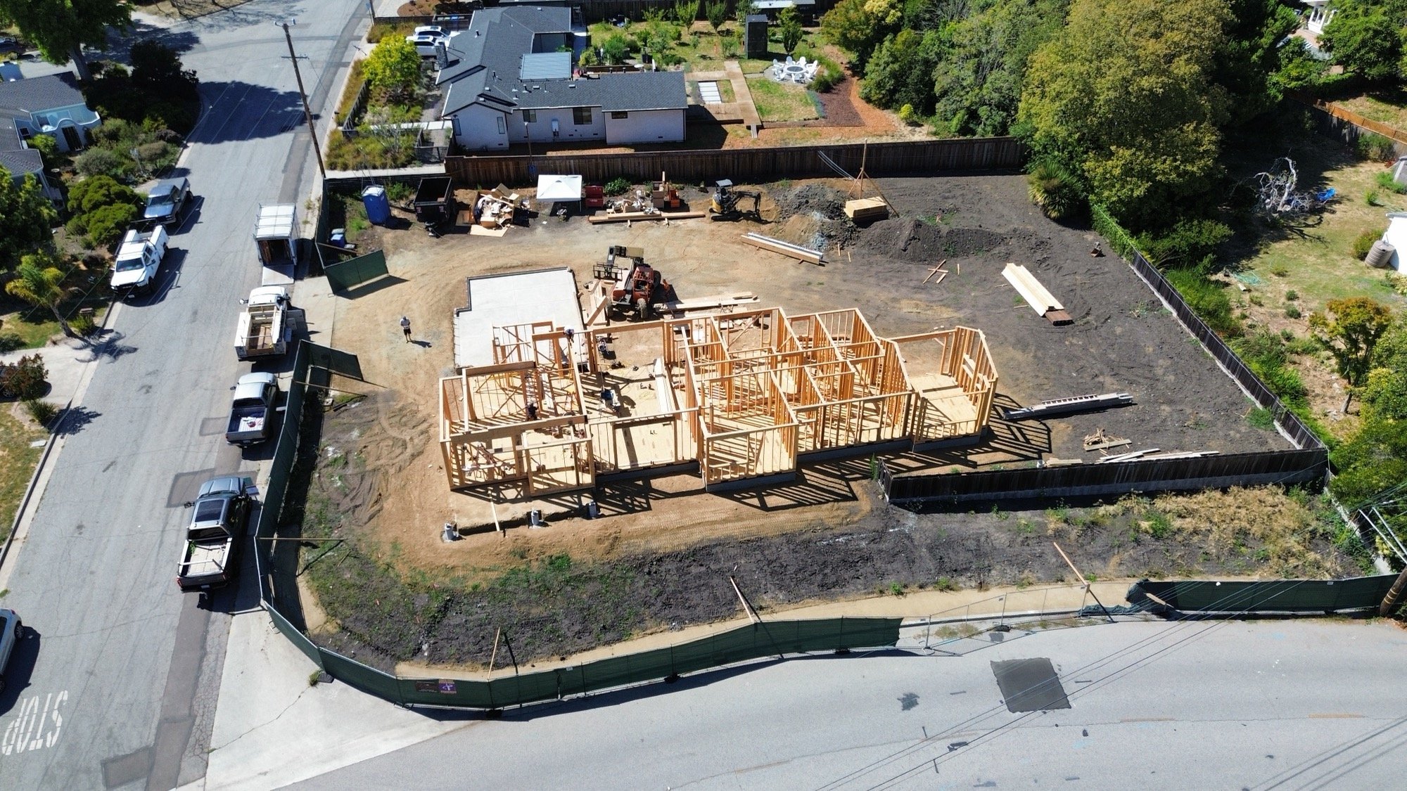 A house under construction with wooden framing in a residential neighborhood, surrounded by a fenced yard, with construction vehicles and materials on site.