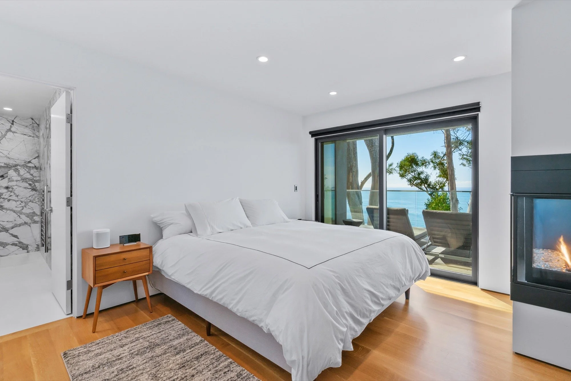 A bedroom with white walls, a bed with white linens, and a sliding glass door opening to an outdoor balcony with chairs and a view of the ocean and trees.