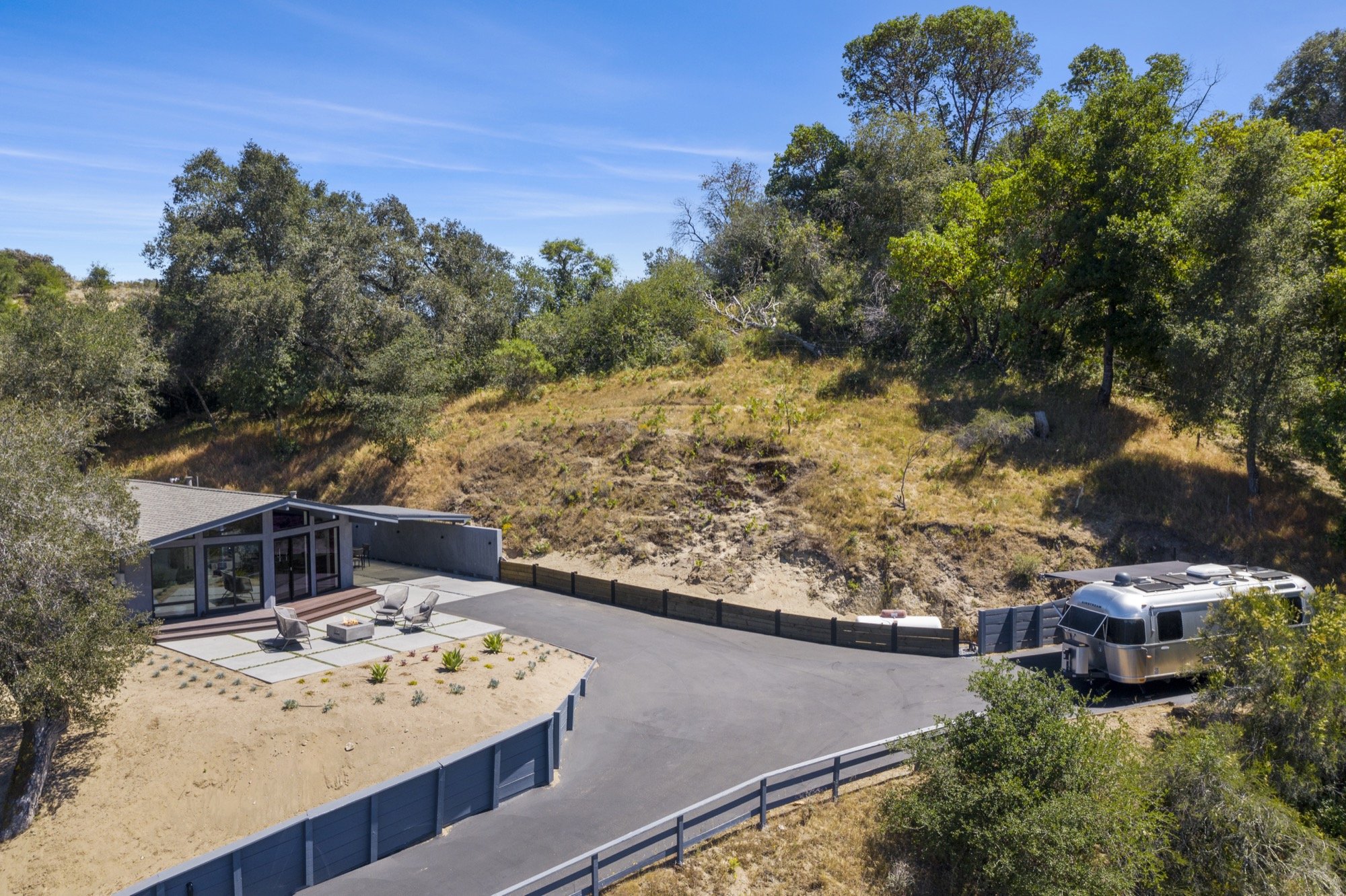 Modern house with large glass windows and outdoor seating area, surrounded by trees and hillside, with the driveway leading to a parked vintage silver camper trailer.