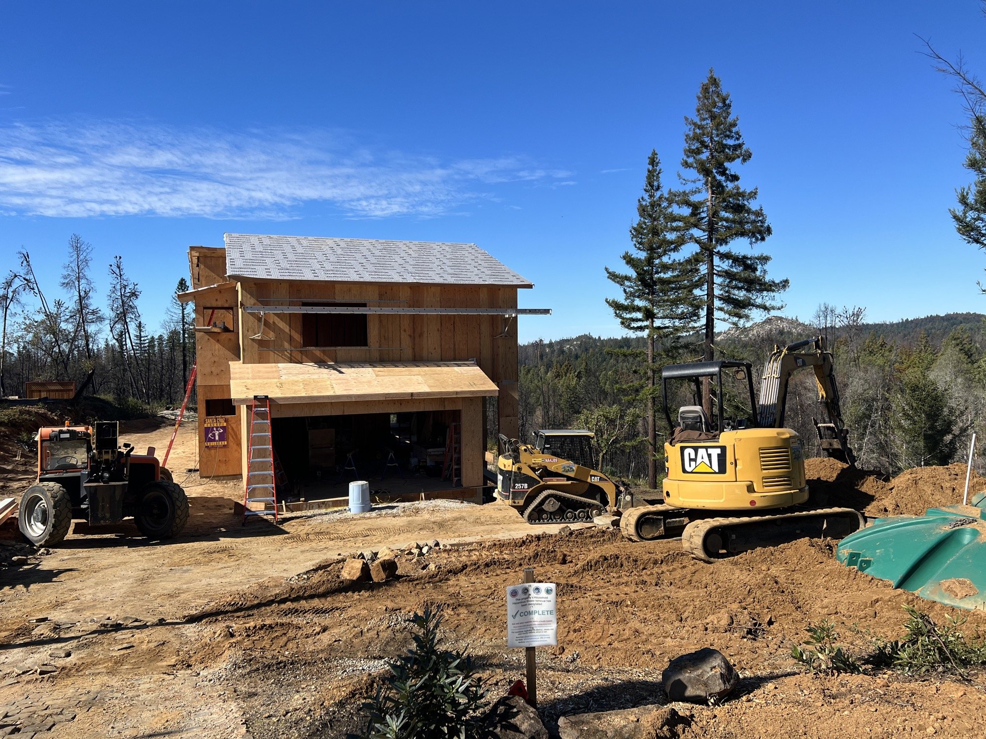Construction site of a wooden house with construction equipment, including a CAT excavator, a mini bulldozer, and a tractor, against a backdrop of blue sky and trees.