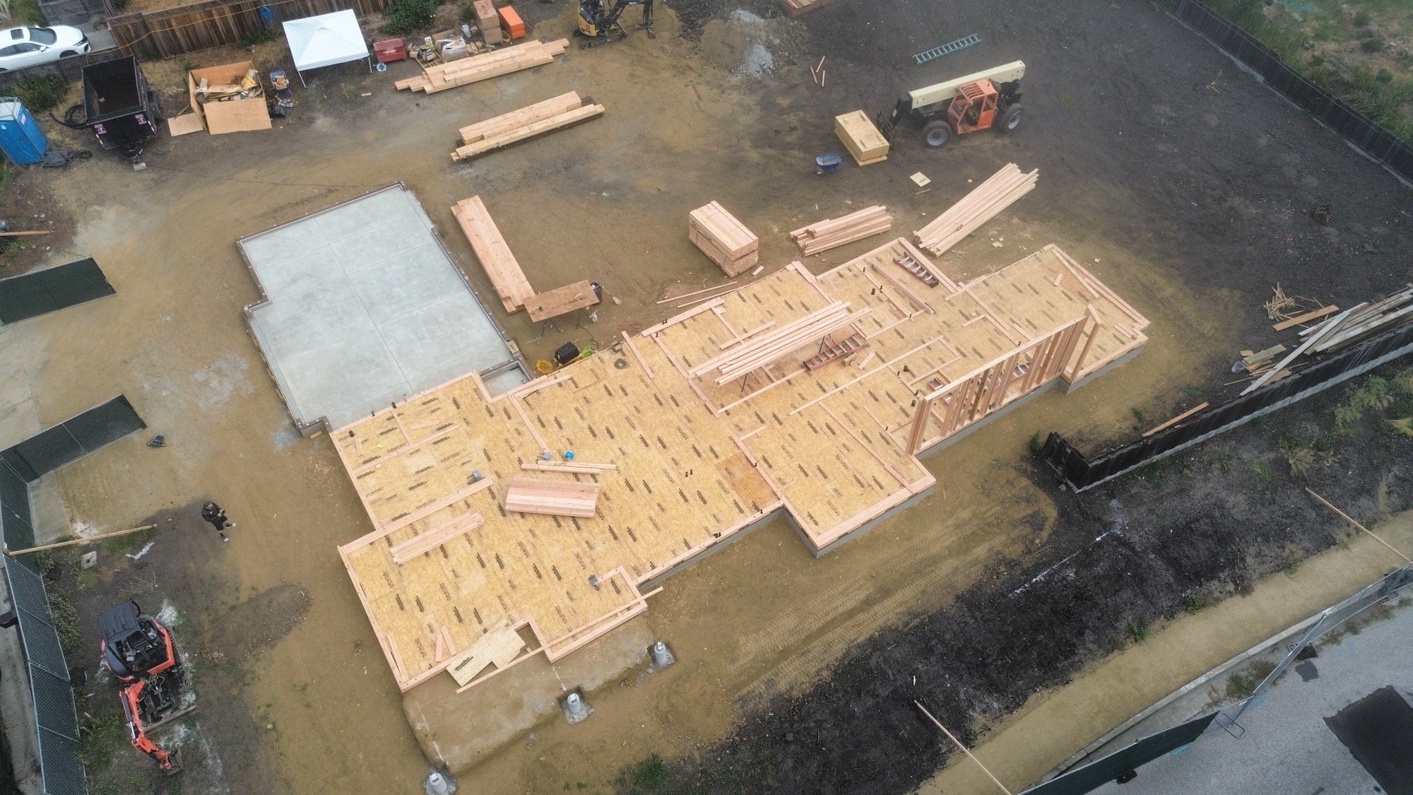 An aerial view of a construction site showing wooden framing, construction materials, and machinery working on building a house, with sections of concrete foundation already poured.