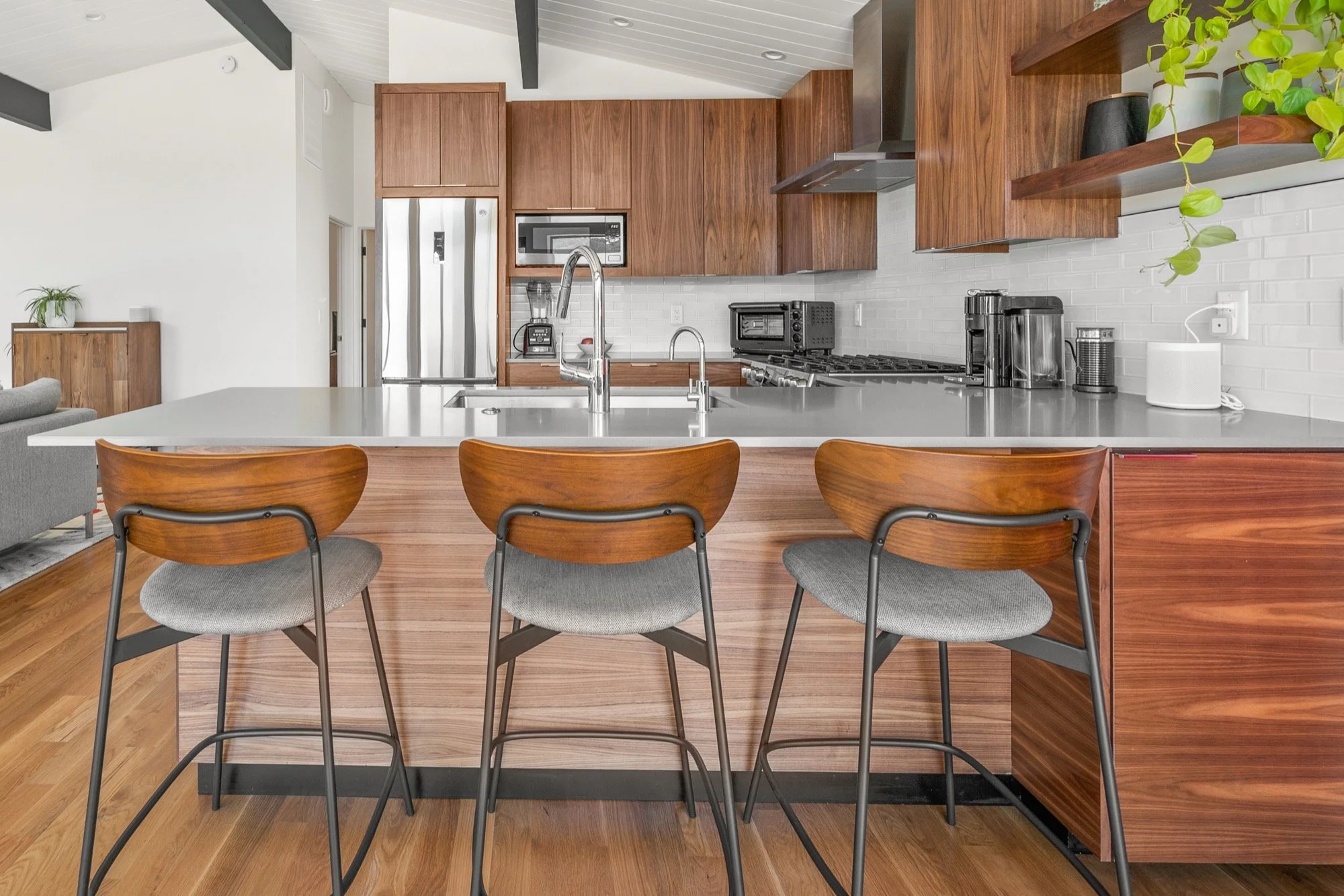 Modern kitchen with wooden cabinets, a white countertop island, three wooden barstools, stainless steel appliances, and open shelving with potted plants.