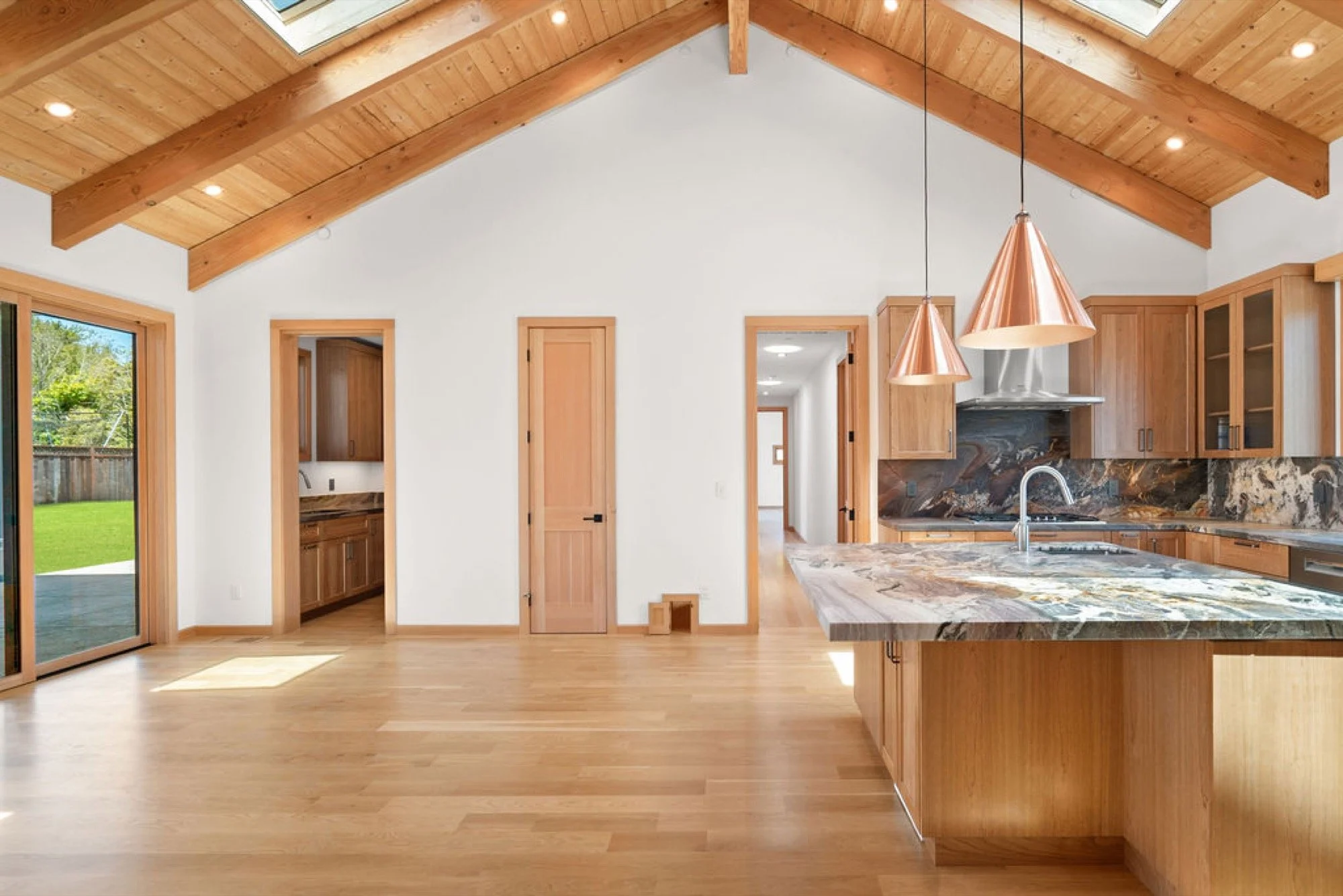 Open kitchen with wooden cabinets, a marble island, copper pendant lights, and a vaulted ceiling with skylights. Sliding door leads to backyard with grass and trees.