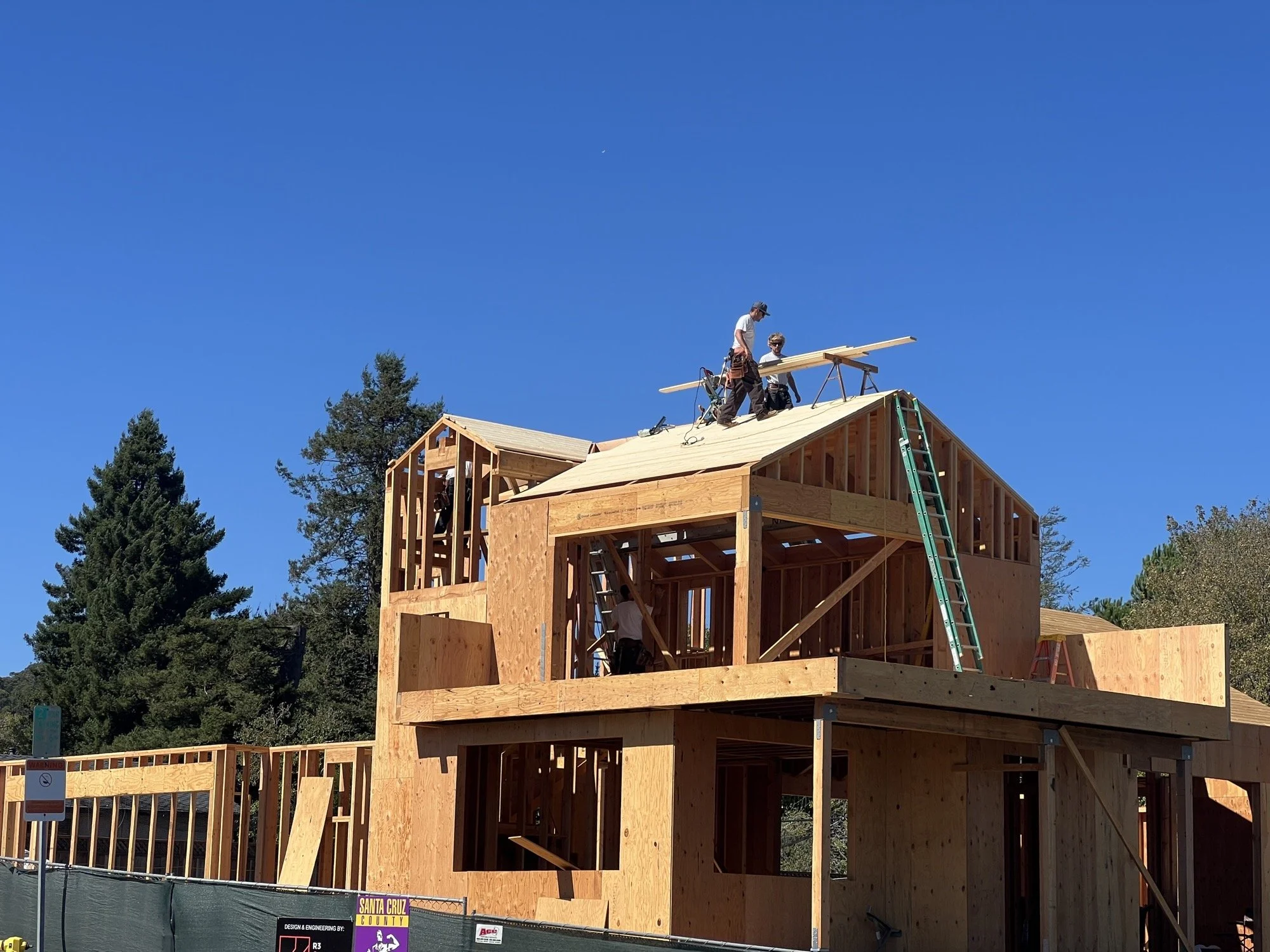 Construction workers building a two-story wooden house on a clear, sunny day with trees in the background.