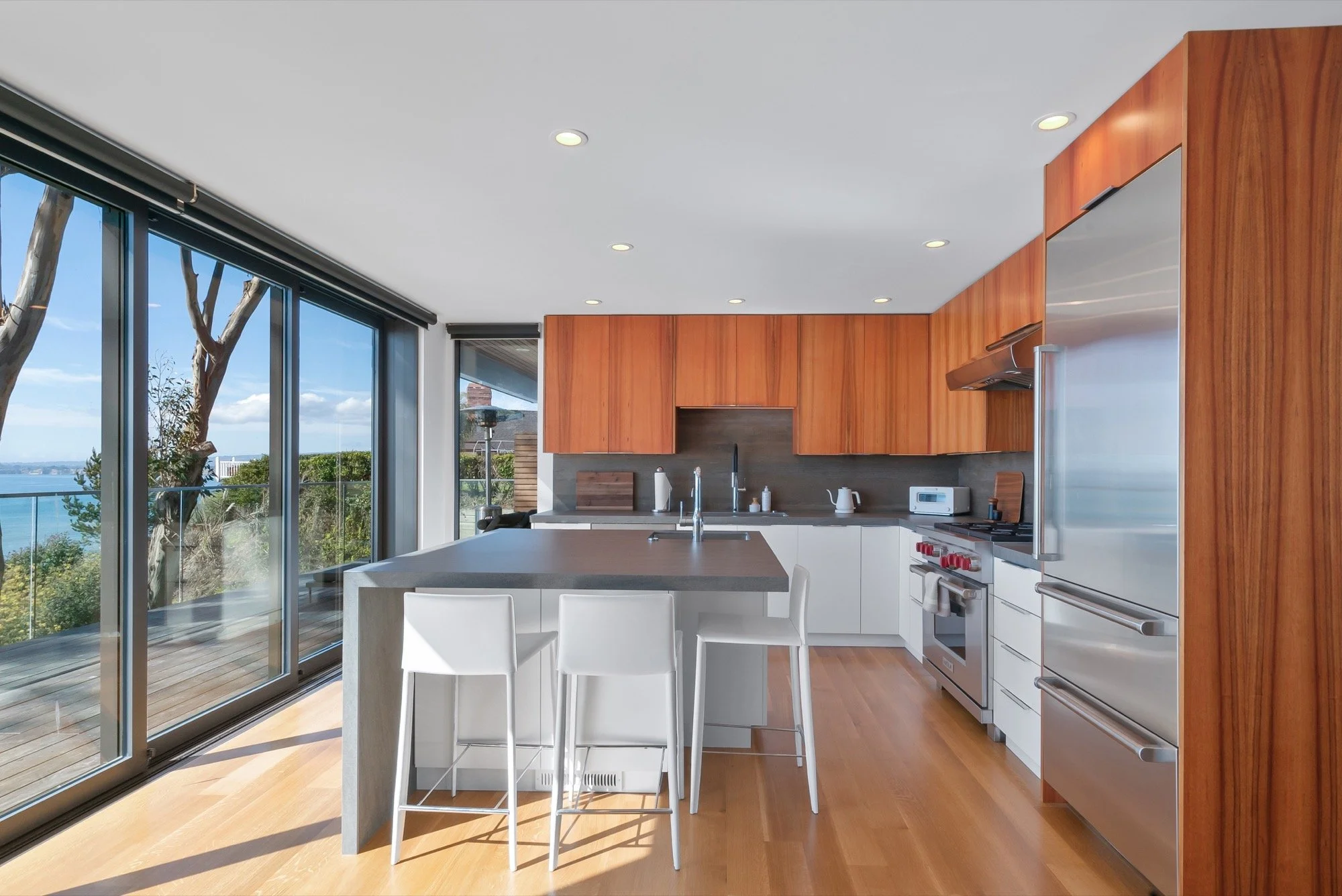 Modern kitchen with large windows overlooking a scenic outdoor view, featuring wooden cabinetry, stainless steel appliances, and a central island with white chairs.
