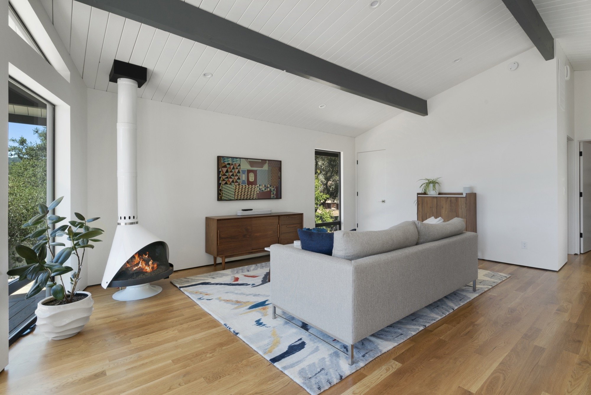 A modern living room with white walls and a wooden floor, featuring a white sofa, a colorful area rug, a wooden sideboard, a wall-mounted TV, a fireplace with a white chimney, and large windows showing greenery outside.