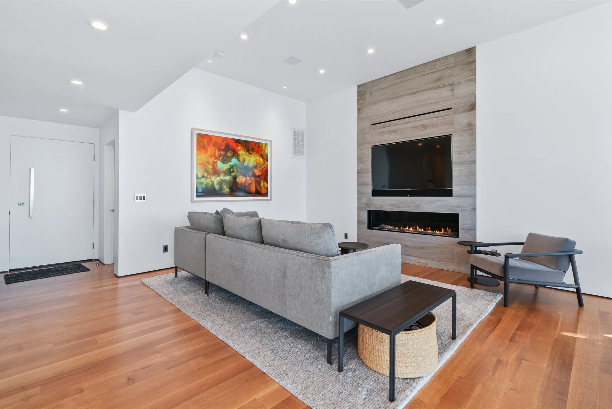 Modern living room with a gray sofa, wood flooring, a wall-mounted fireplace, a large flat-screen TV, an abstract colorful painting, and a gray armchair.