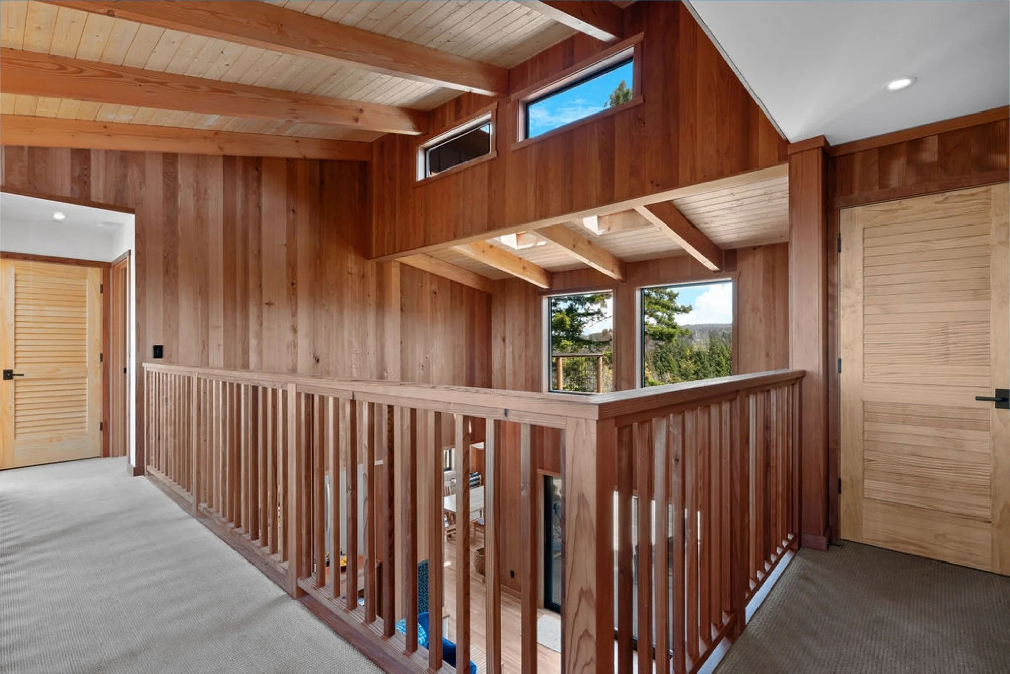 Wood-paneled interior of a home with high ceiling, large windows, wooden doors, and a staircase rail leading downstairs.