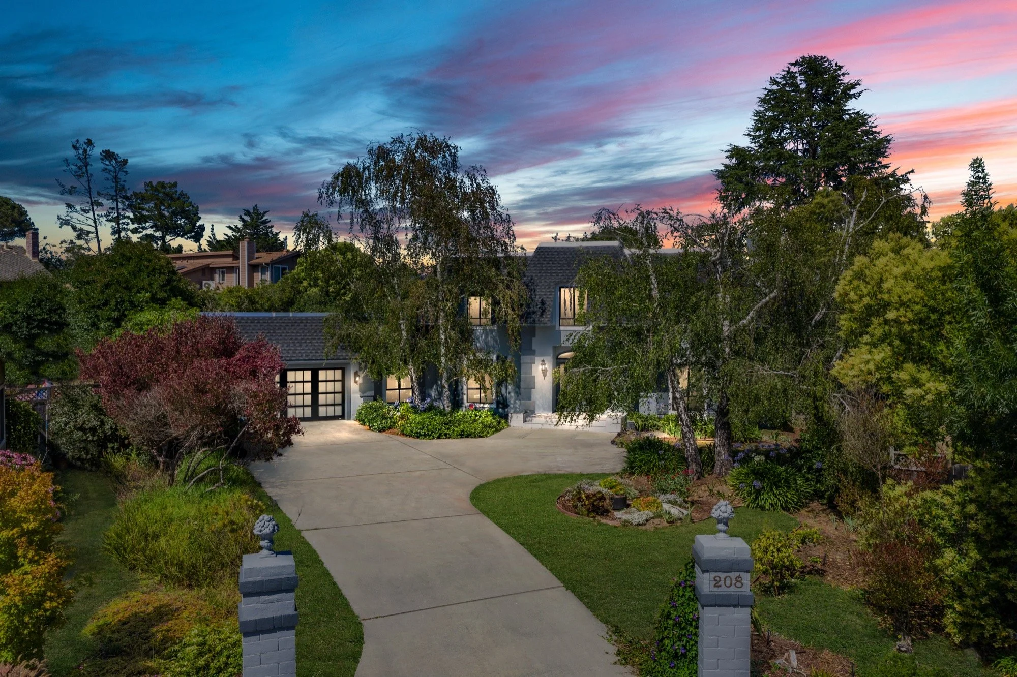 A two-story modern house with a gray exterior, black-framed windows, a front door, and a garage. The house is surrounded by well-maintained landscaped garden with various trees and shrubs. The driveway and walkway are concrete, and the scene is set at dusk with a colorful sky.