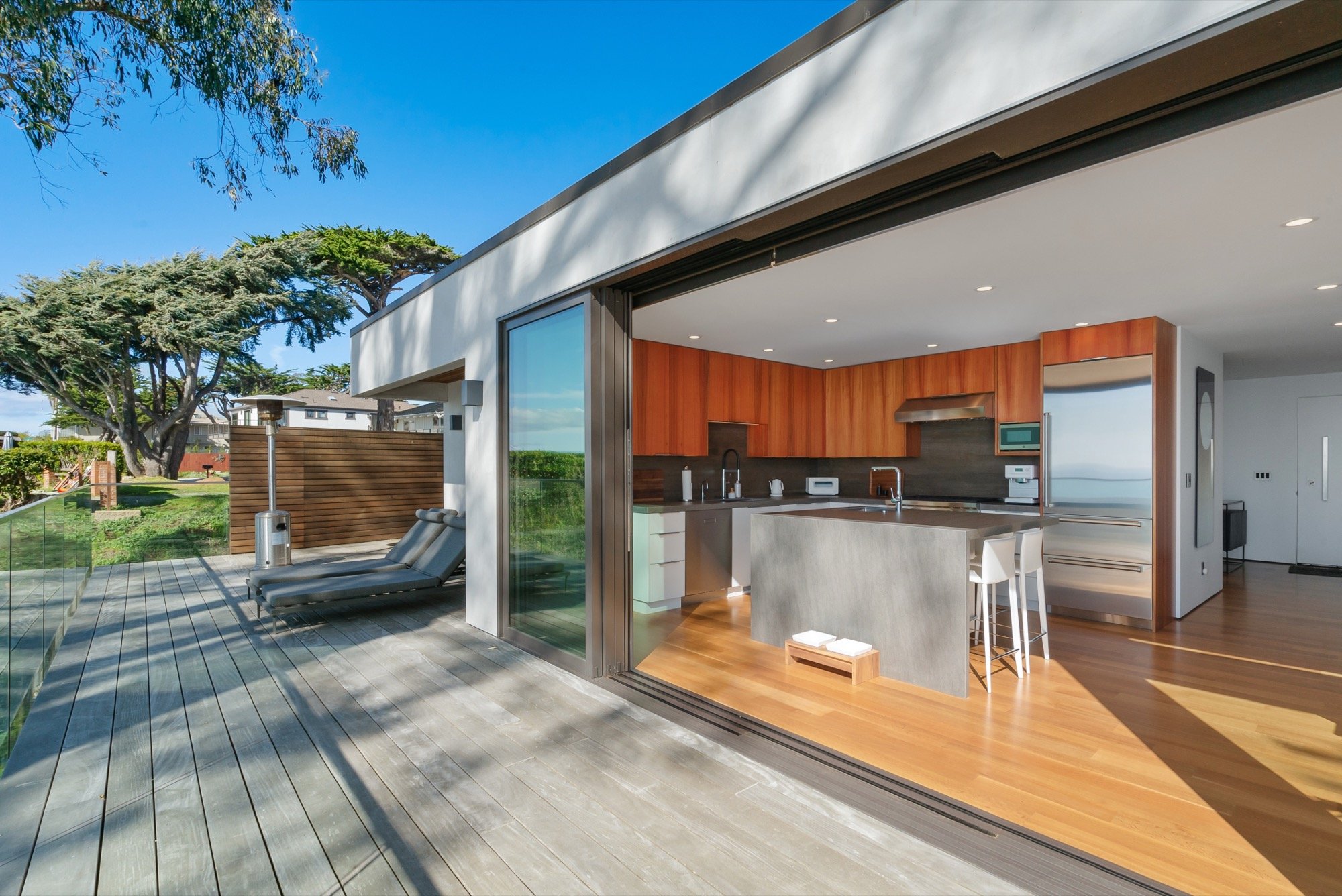 Modern outdoor wooden deck leading to an open indoor kitchen with wooden cabinets, stainless steel appliances, and a gray kitchen island, with lounge chairs, trees, and a clear blue sky in the background.