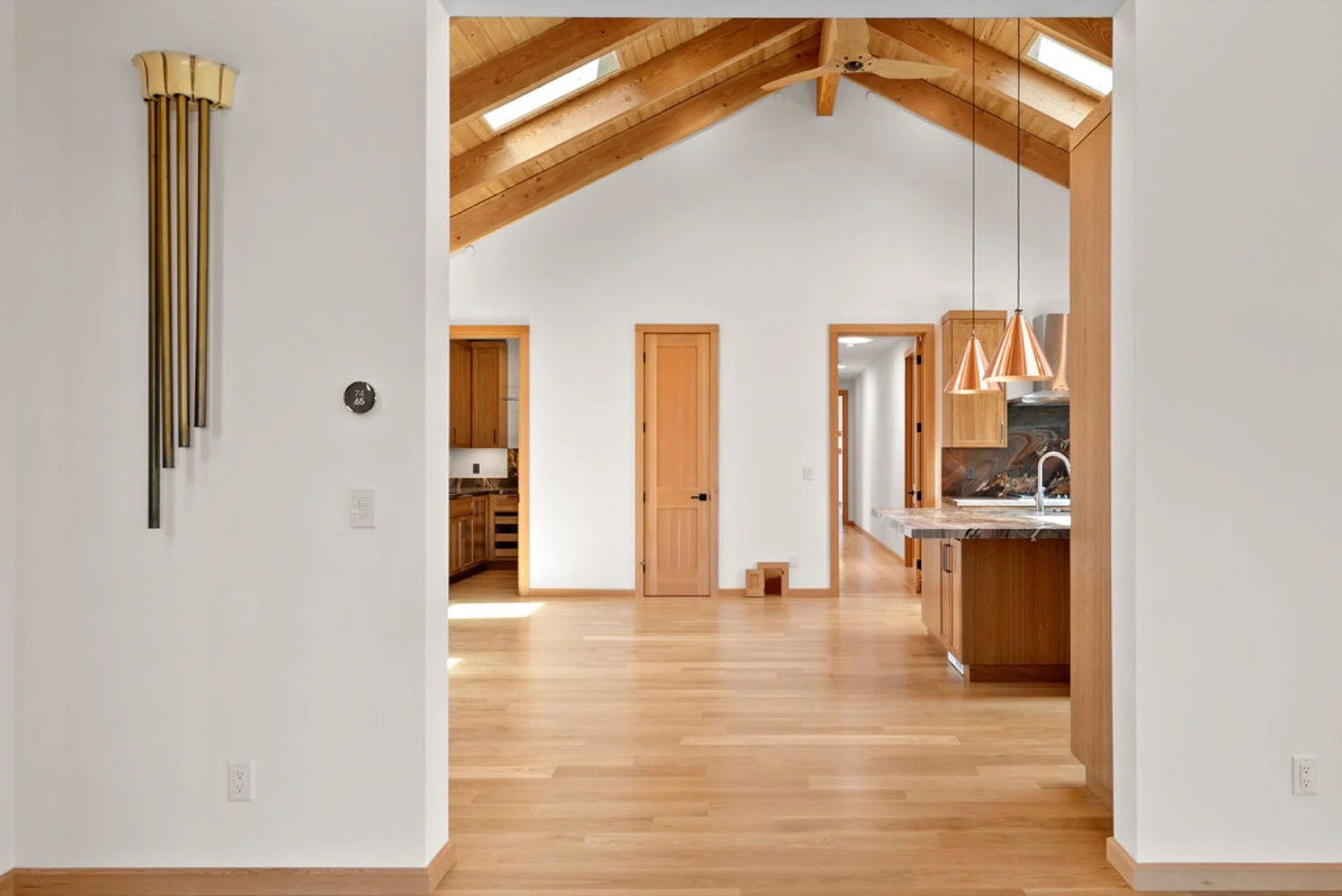 Interior view of a house with high vaulted wooden ceilings and skylights, featuring a kitchen with wooden cabinets, a kitchen island with a marble countertop, and a hallway leading to other rooms, with natural wood flooring and trim.