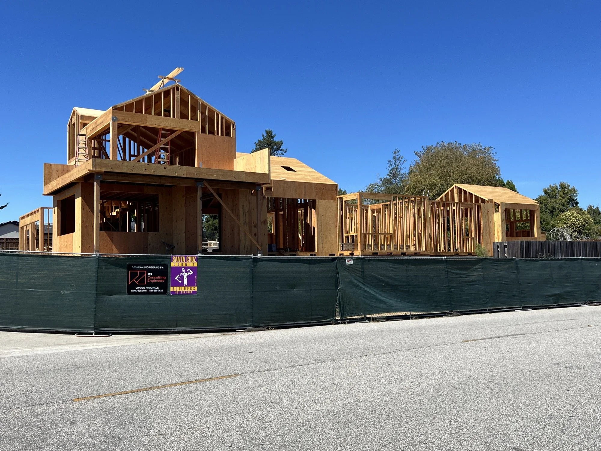 Wooden house under construction with framing, scaffolding, and a green construction fence in front, under a clear blue sky.