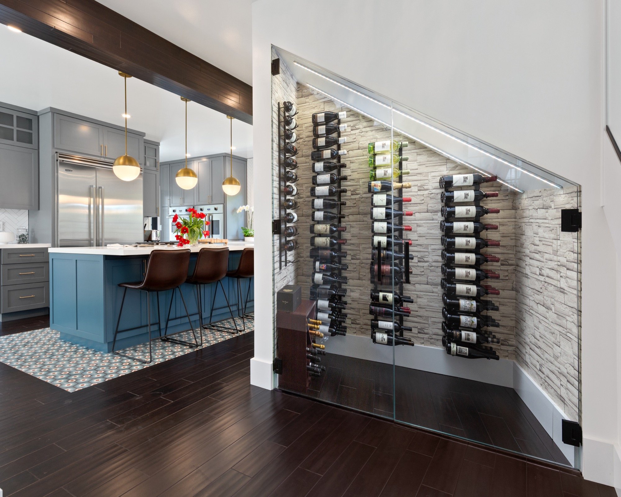 Modern kitchen with island, pendant lighting, blue cabinets, bar stools, floral arrangement, and a wine storage wall with bottles on a sloped wall with a glass door.