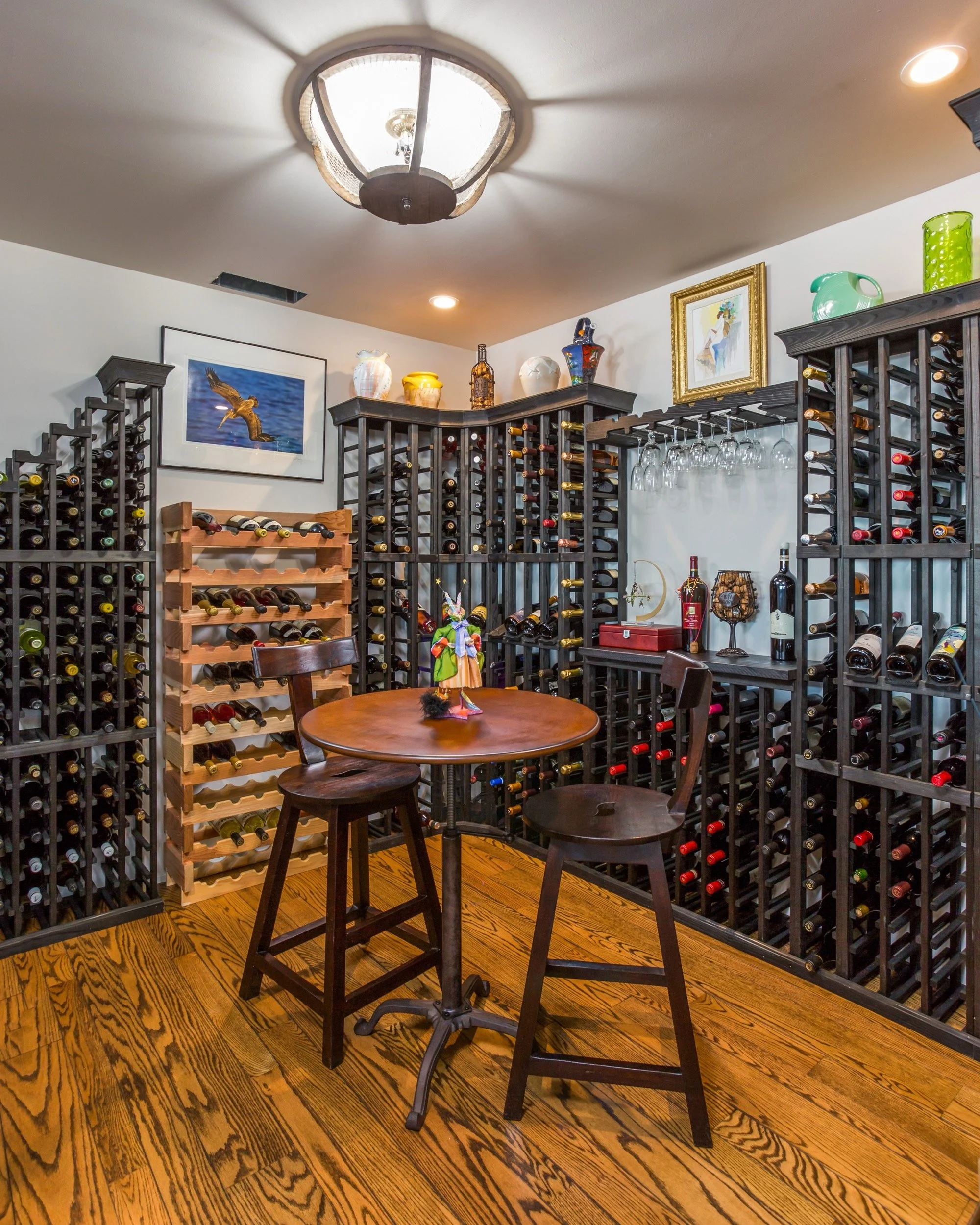 Wine cellar with wooden and black wine racks filled with bottles, a small round table with two chairs, decorative artifacts on top of the racks, and framed pictures on the wall.