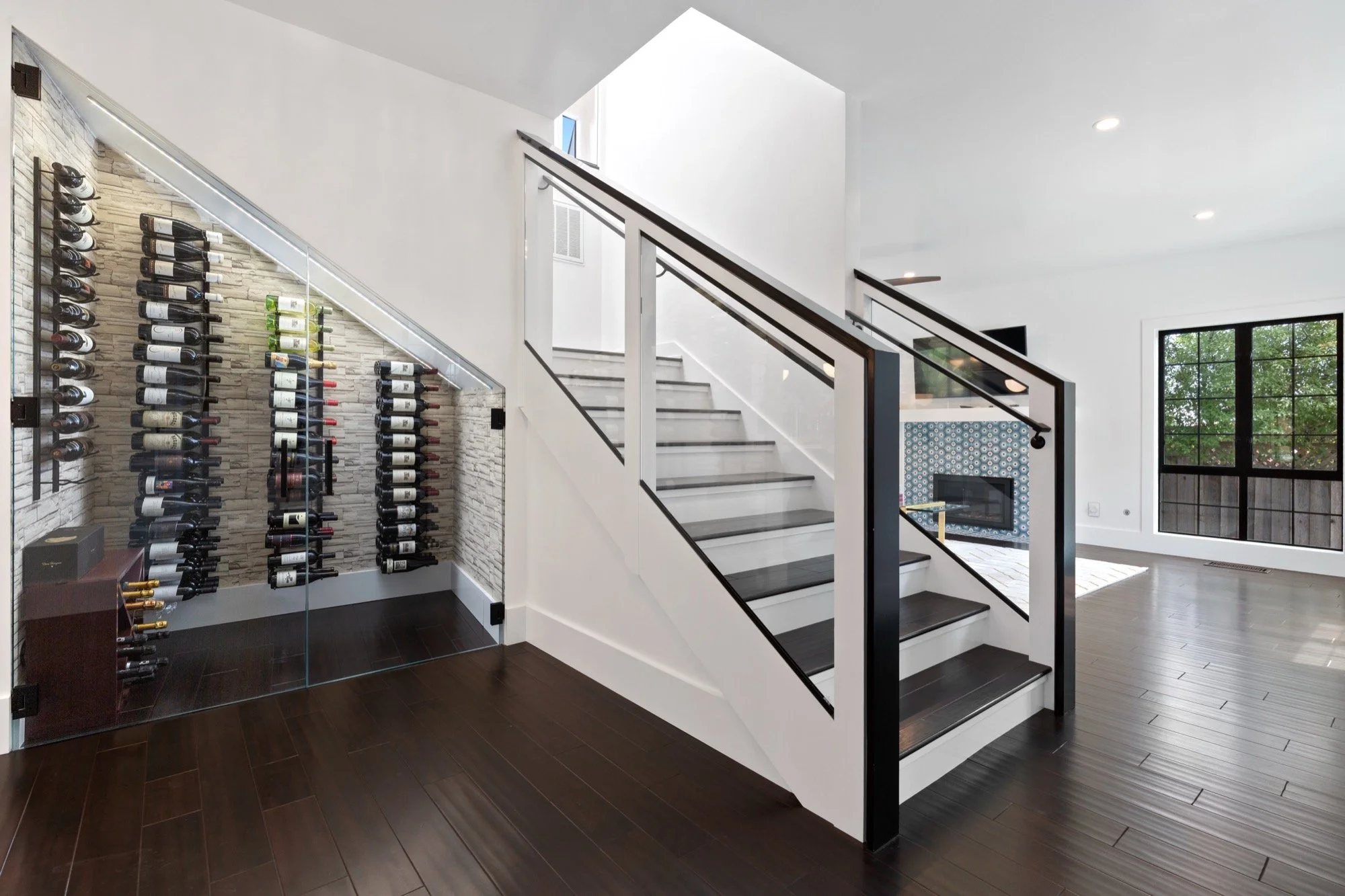 Interior of a modern home with a staircase, a glass-enclosed wine rack, and a fireplace with patterned tile wall, large window, and wood flooring.