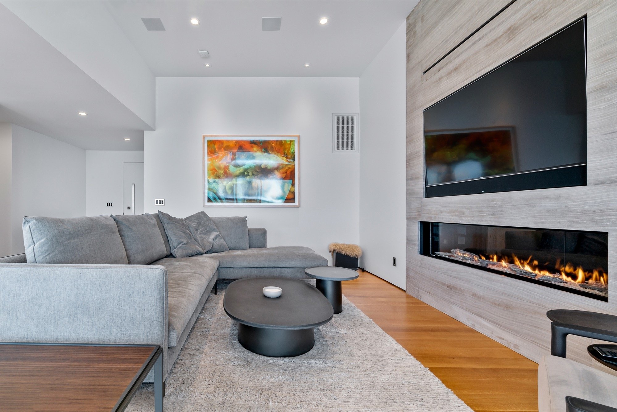 Modern living room with gray sectional sofa, abstract art on white wall, wood-paneled fireplace with a television above, hardwood floor, and a beige area rug.
