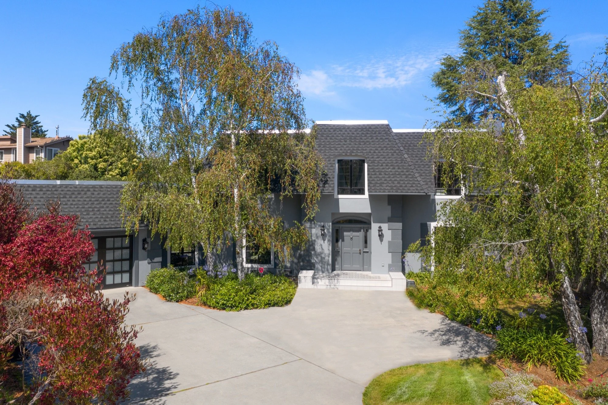 Gray two-story house with front steps, surrounded by trees and colorful shrubs, and a driveway leading to a garage on the left.