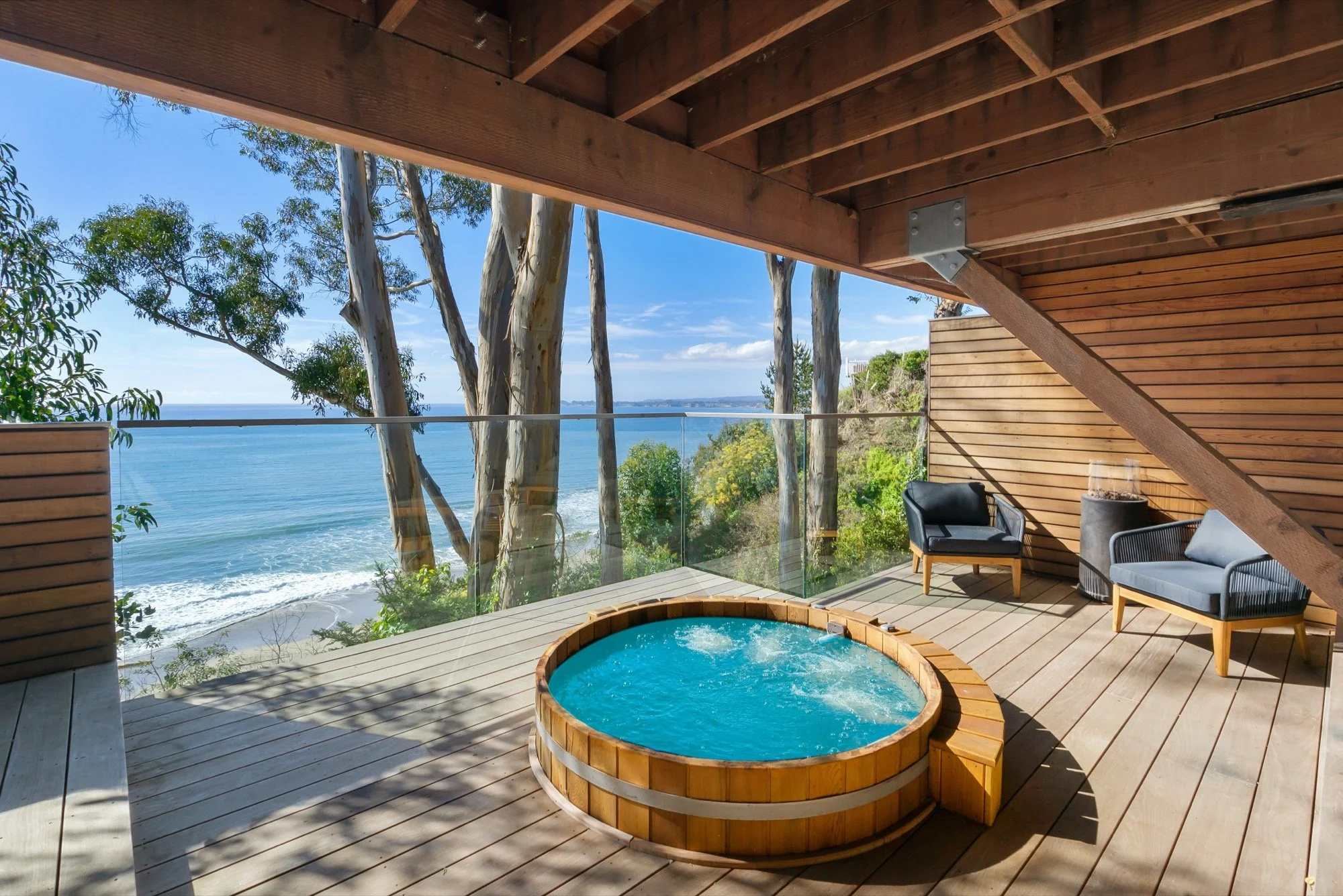 A wooden deck overlooking the ocean with a hot tub, two modern outdoor chairs, and tall trees nearby, under a partially open roof.