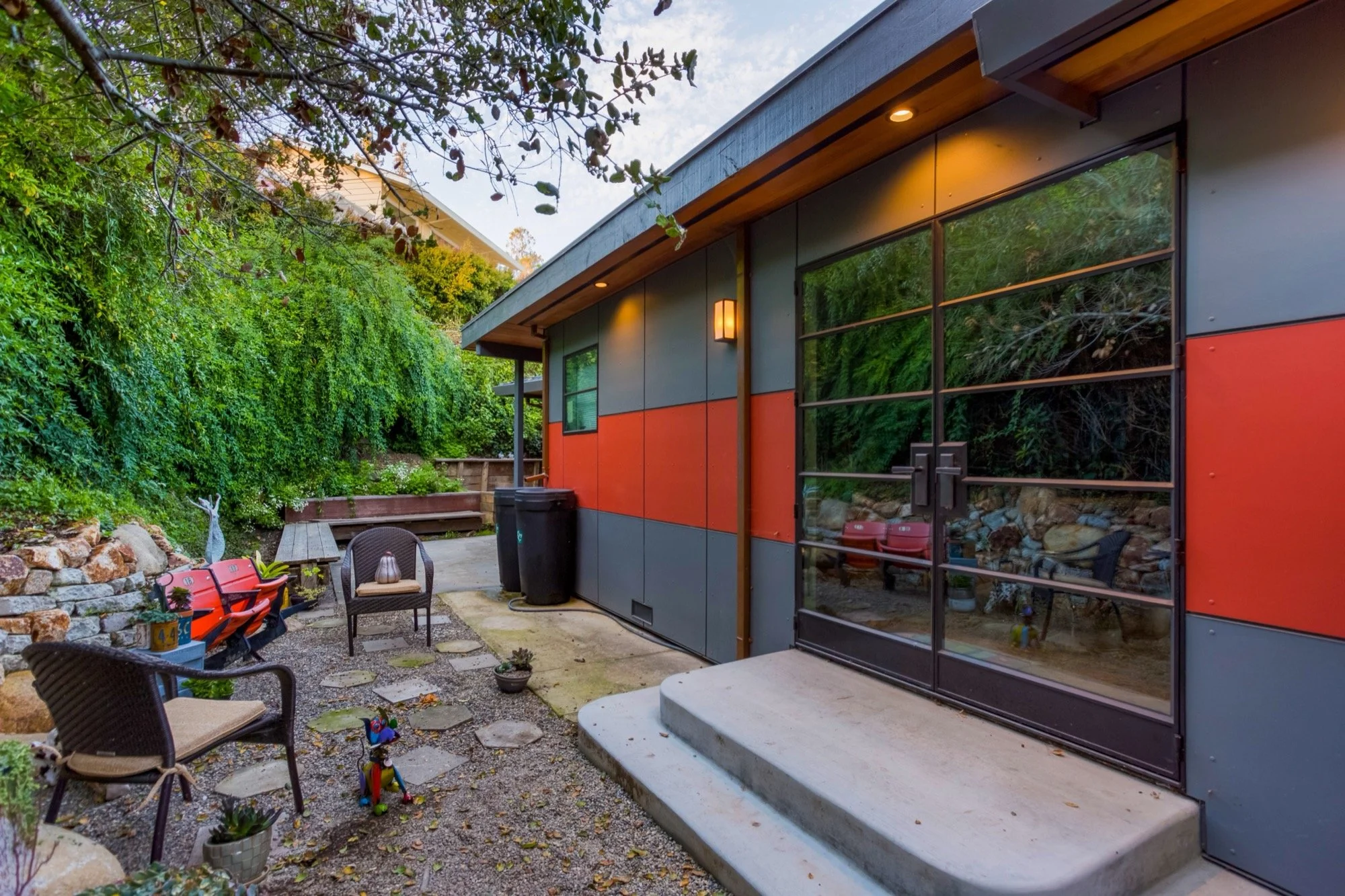 A modern backyard patio area with outdoor seating, a small toy dinosaur, potted plants, and a house with sliding glass doors and colorful exterior walls, surrounded by lush green foliage.