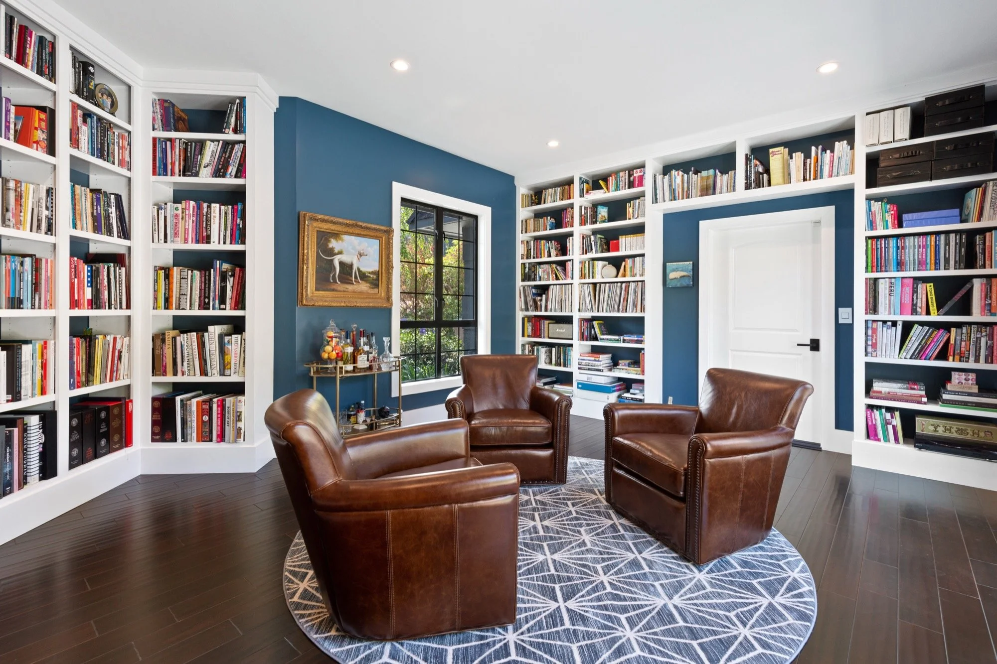 A cozy reading corner with four brown leather armchairs arranged in a circle on a geometric-patterned rug, surrounded by floor-to-ceiling white bookshelves filled with colorful books, a window with a view of outdoor greenery, blue accent walls, and a