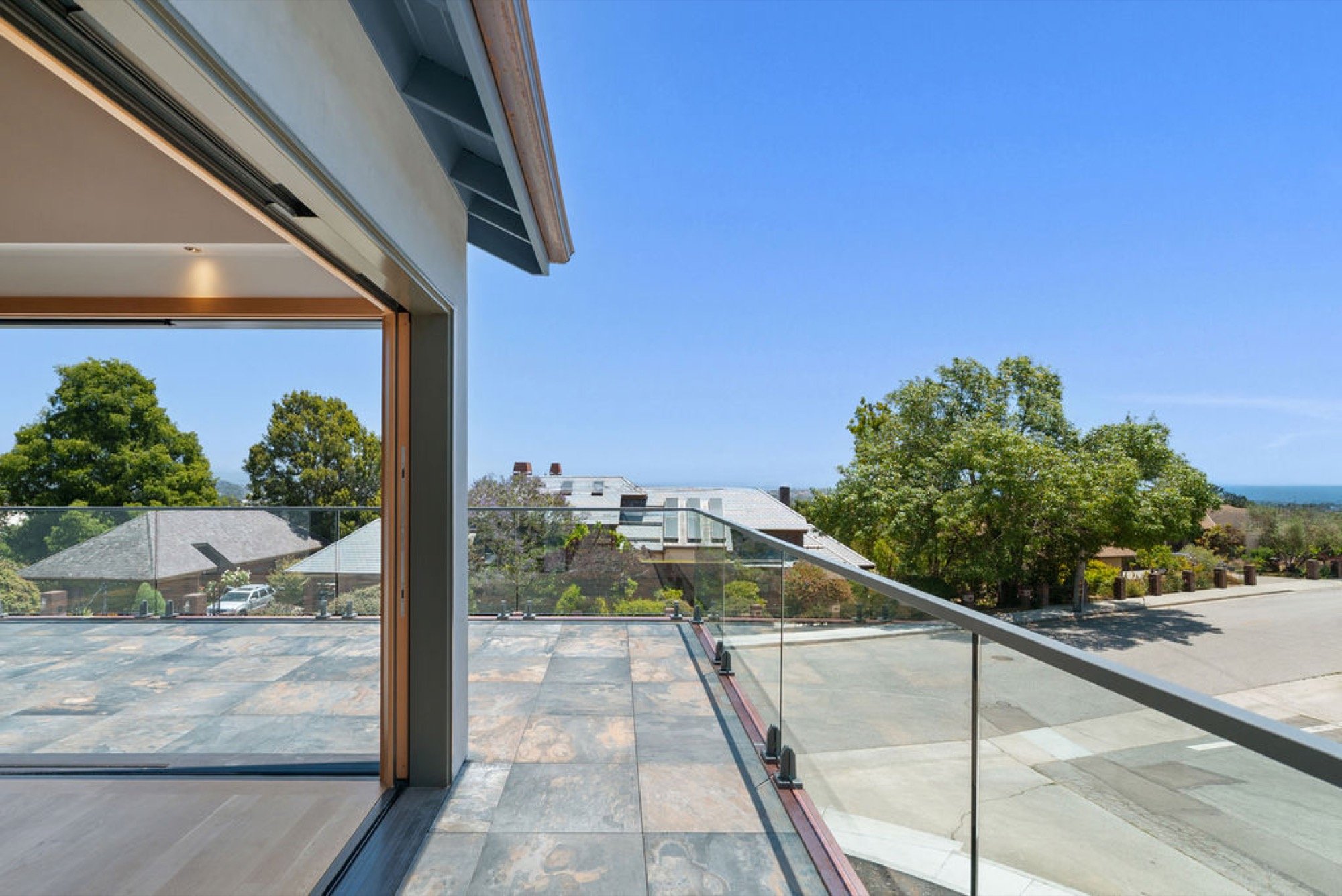 View from a balcony with glass railing overlooking a neighborhood with trees and houses under a clear blue sky.