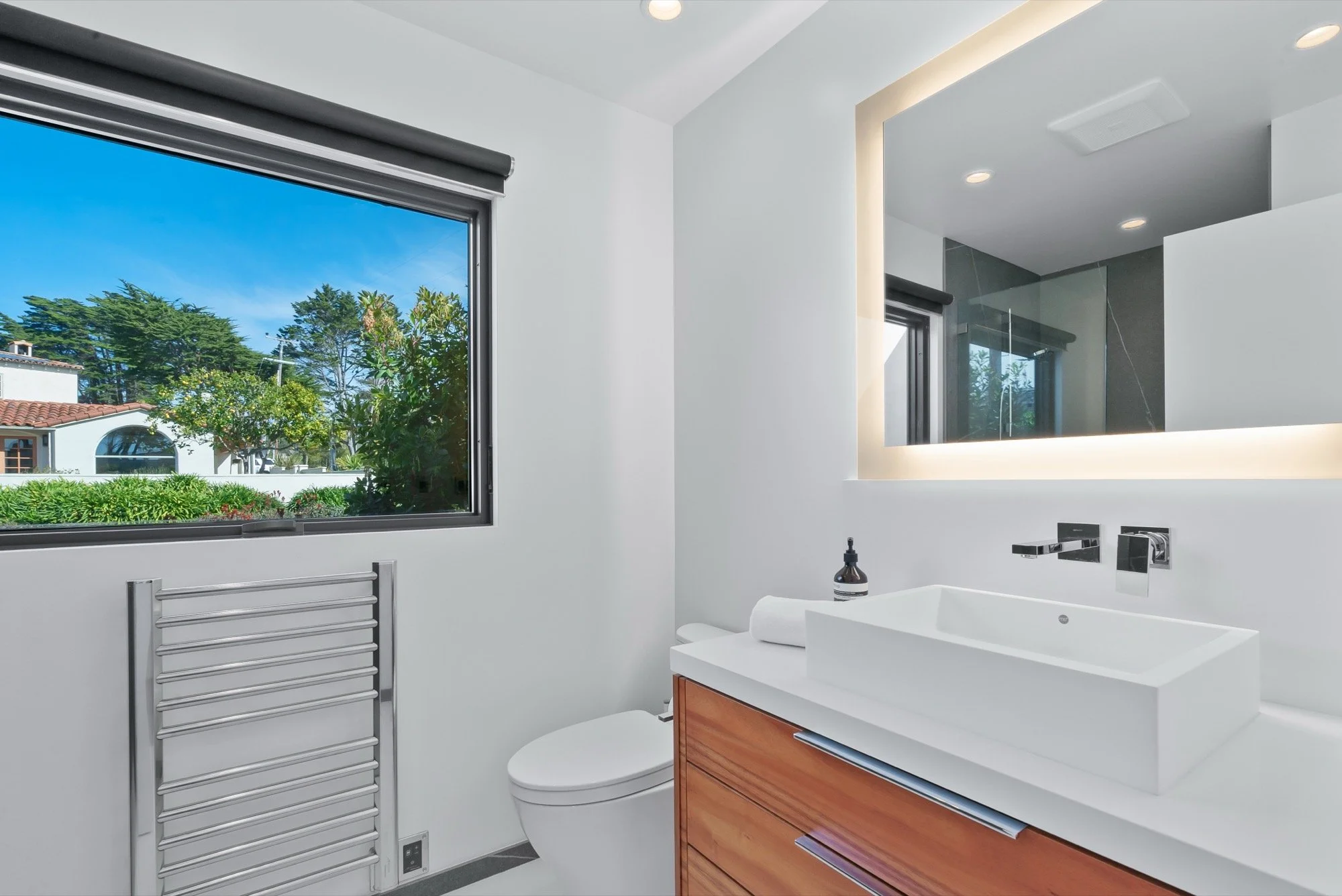 Modern bathroom with white walls, large window showing greenery outside, wooden cabinet with a white vessel sink, mirror with backlit LED lighting, and a towel radiator.