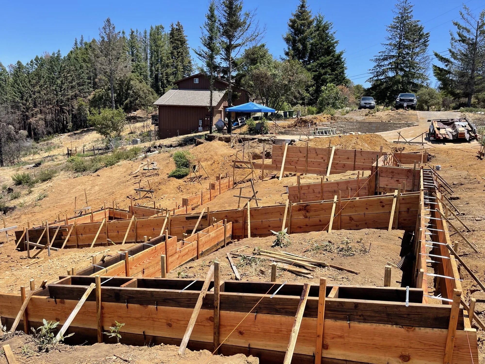 Construction site with wooden forms for building foundations on a sloped land, surrounded by trees and a house in the background.