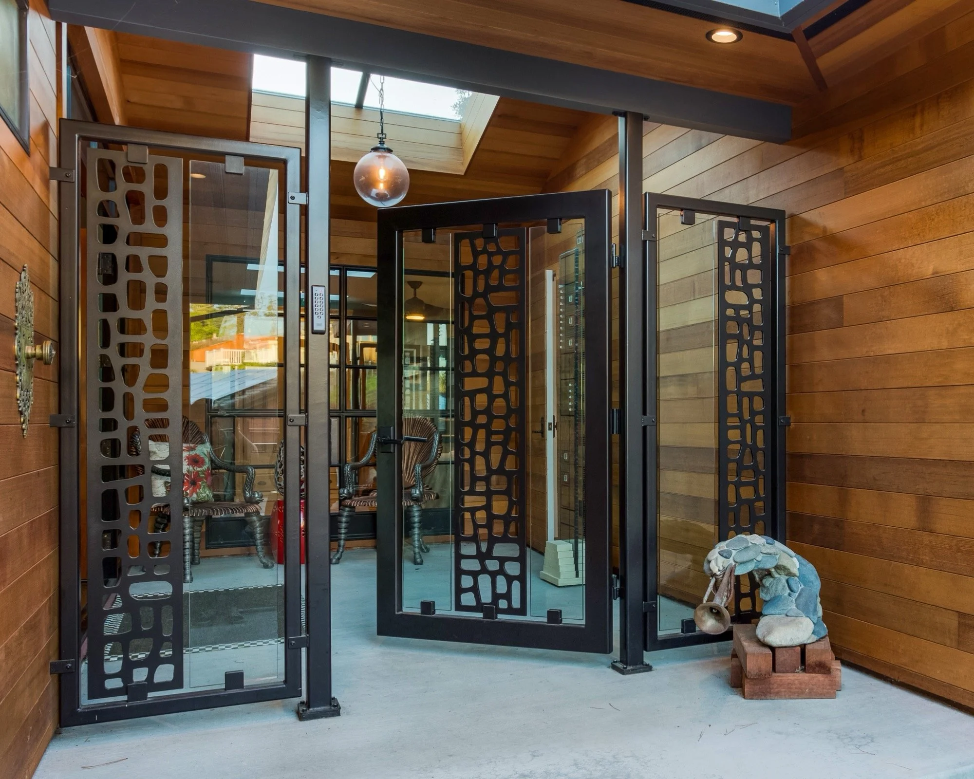 A screened porch with wooden walls, black metal framing, and a glass door, featuring chairs and a hanging light fixture.
