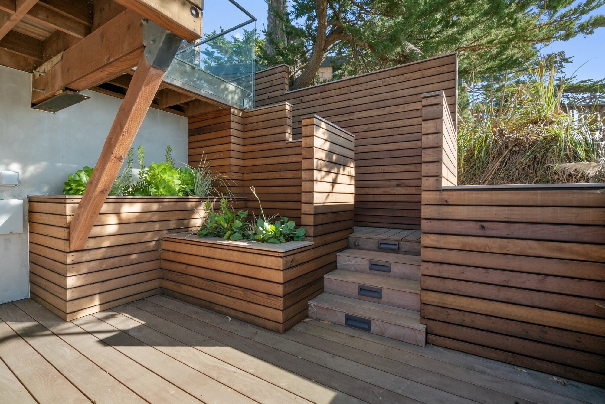 Outdoor wooden deck with built-in planters containing green plants, stairs, and a glass railing, with trees in the background.
