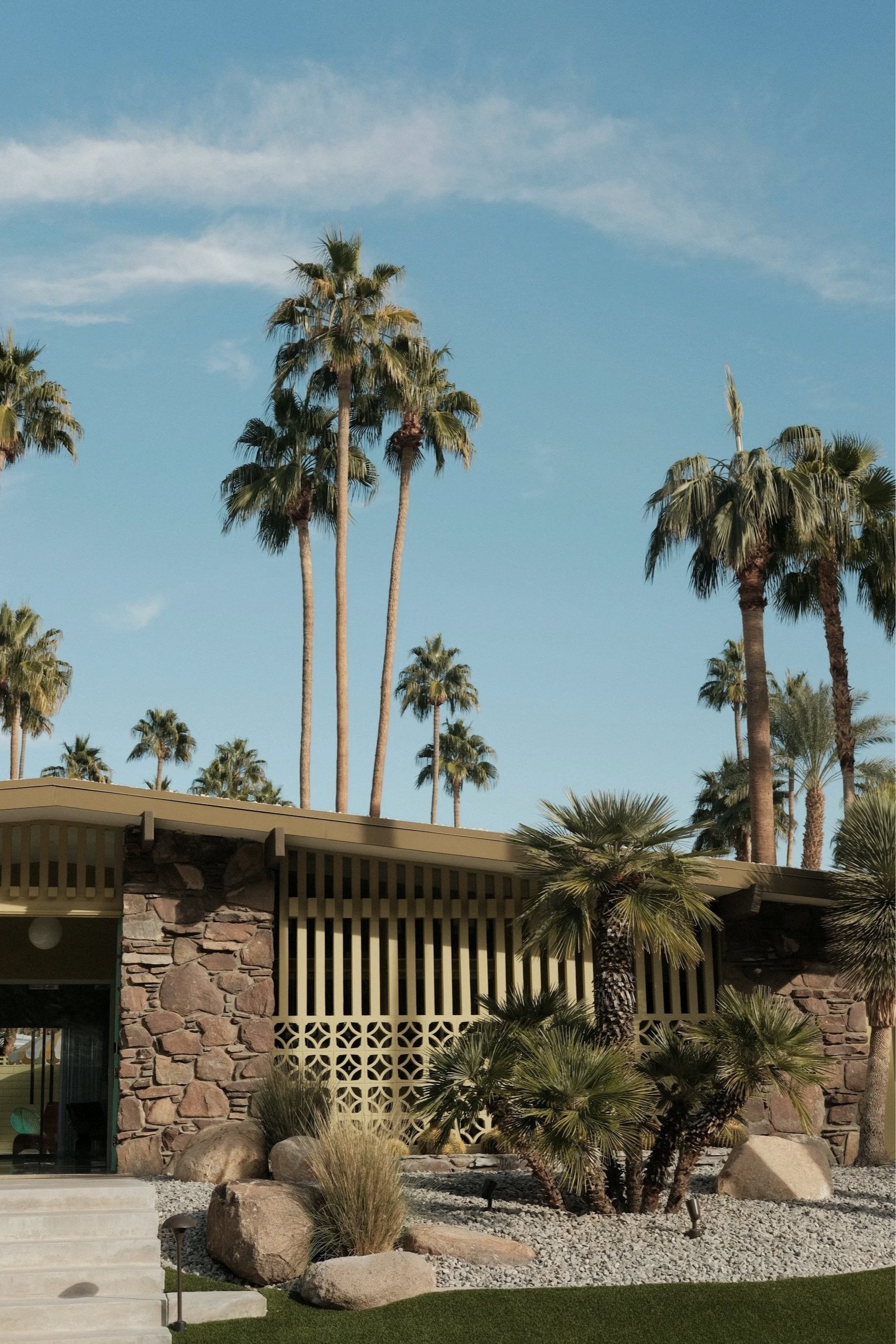 Mid-century modern house with stone and wood exterior, surrounded by desert landscaping and tall palm trees under a blue sky.