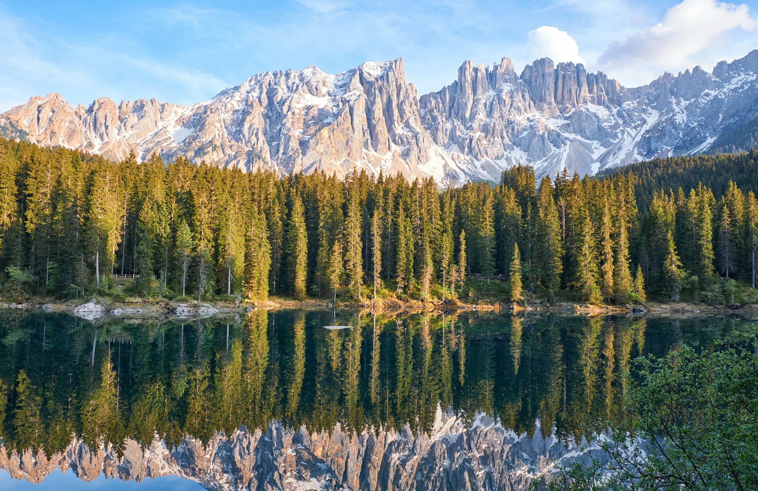 Scenic mountain landscape with snow-capped peaks, dense evergreen forest, and a mirror-like lake reflecting the mountains and trees.