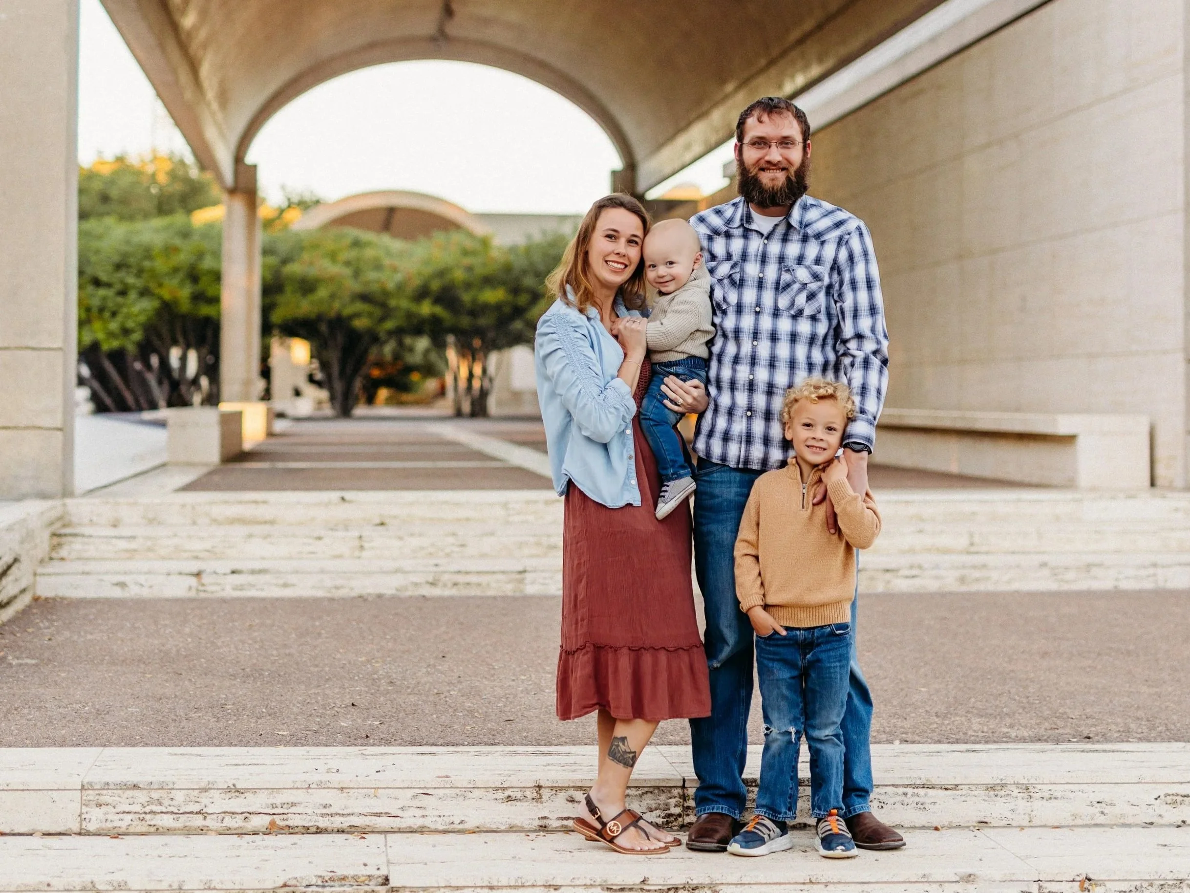 Happy family of four with two children standing outdoors on steps under a concrete arch, smiling at the camera at sunset.