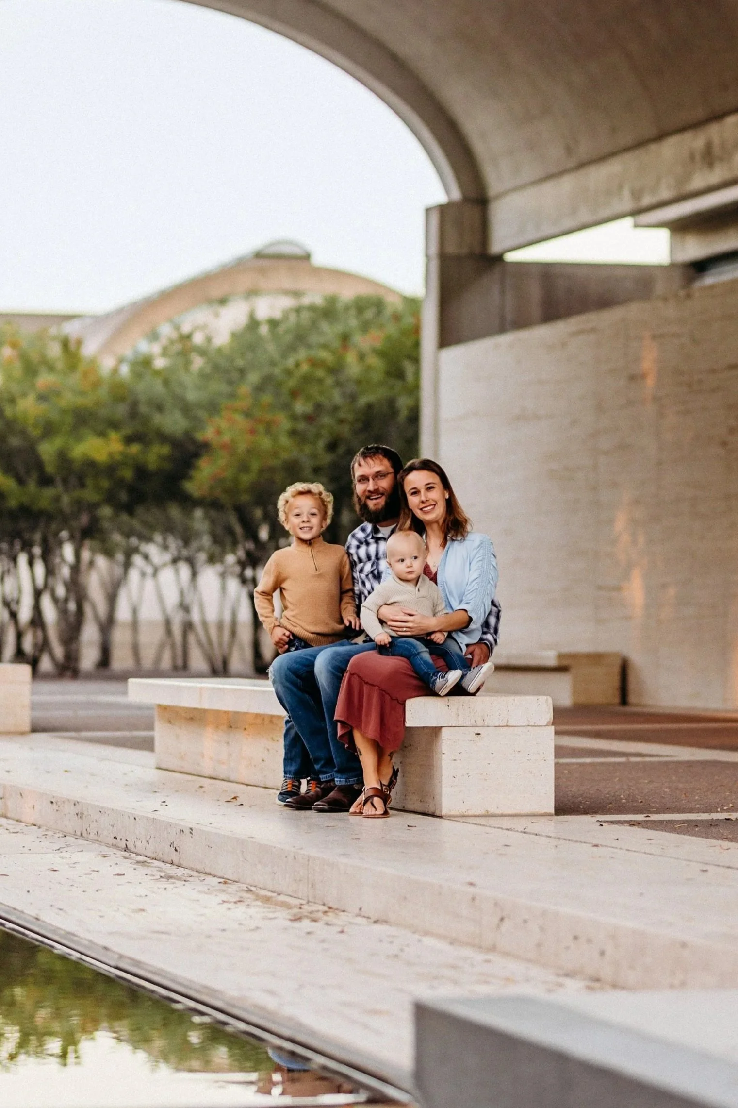 A family of four sitting on a concrete bench outdoors under a bridge, with trees and a large building in the background.