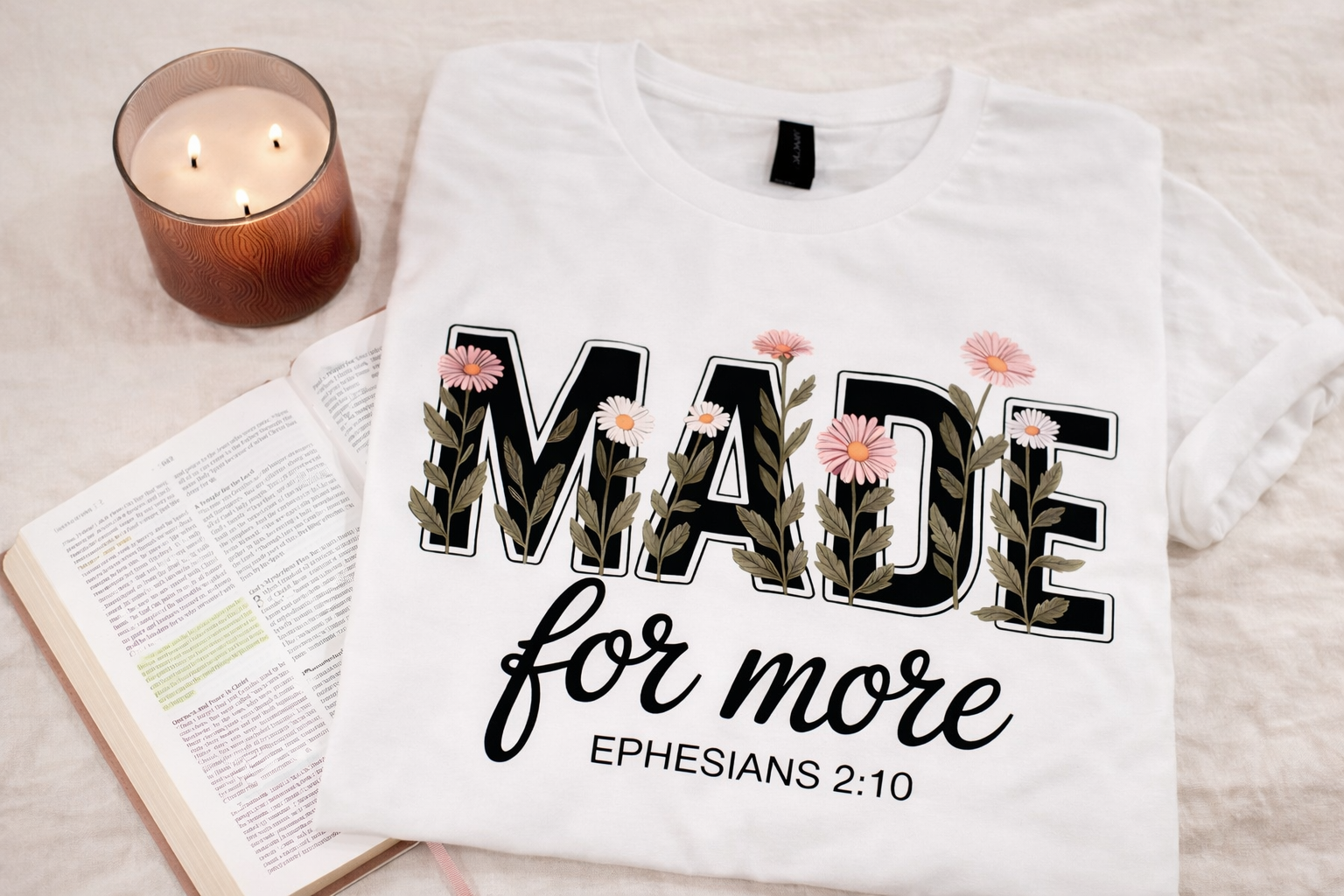 A white t-shirt with the text 'Made for more' and a Bible verse reference 'Ephesians 2:10', decorated with pink flowers and green leaves, laid on a table beside an open Bible and a lit candle in a brown holder.