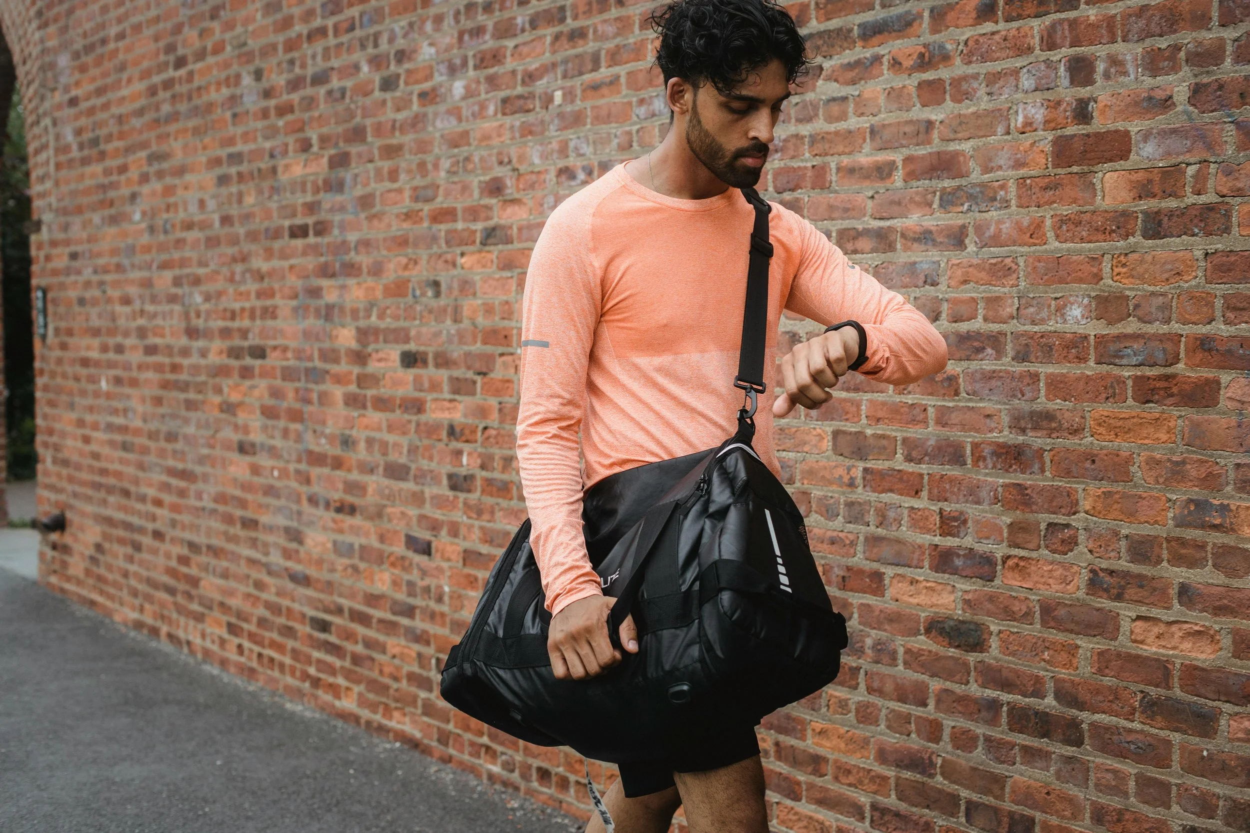 A man in a peach long-sleeve shirt and shorts walking outside next to a brick wall, holding a black gym bag and checking his smartwatch.