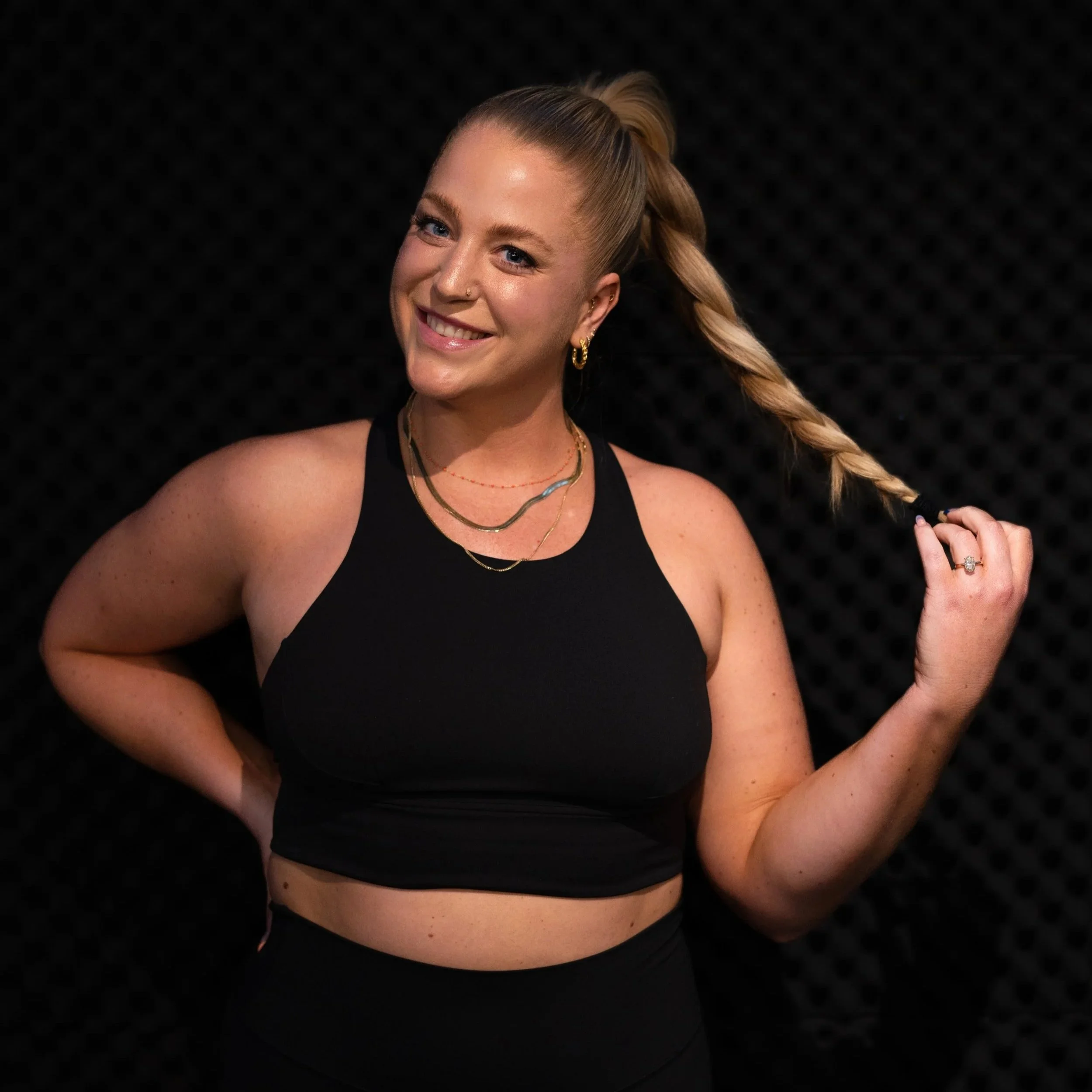 A woman with blonde hair in a long braid, wearing a black crop top, gold earrings, layered necklaces, and smiling while holding her braid in her right hand, against a black textured background.