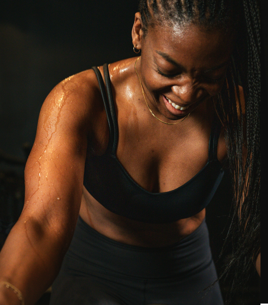 A woman with braided hair and dark skin, sweating and smiling, wearing a black sports bra and black leggings.